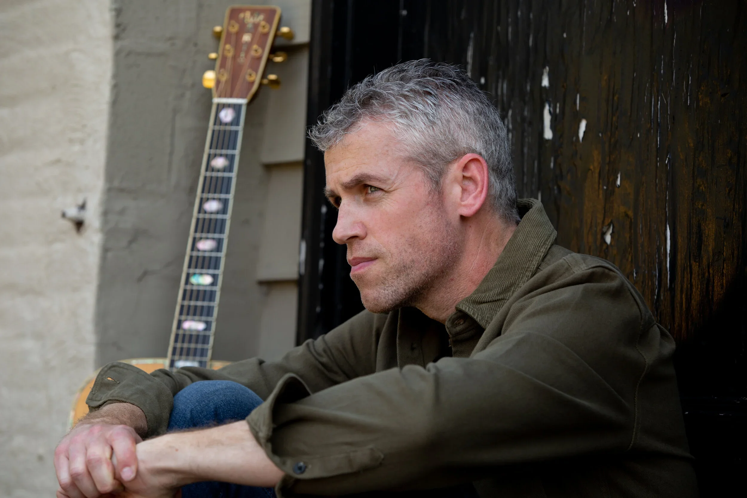 A man with gray hair and a beard sitting outdoors near a dark wooden fence, with a guitar leaning against the wall behind him.
