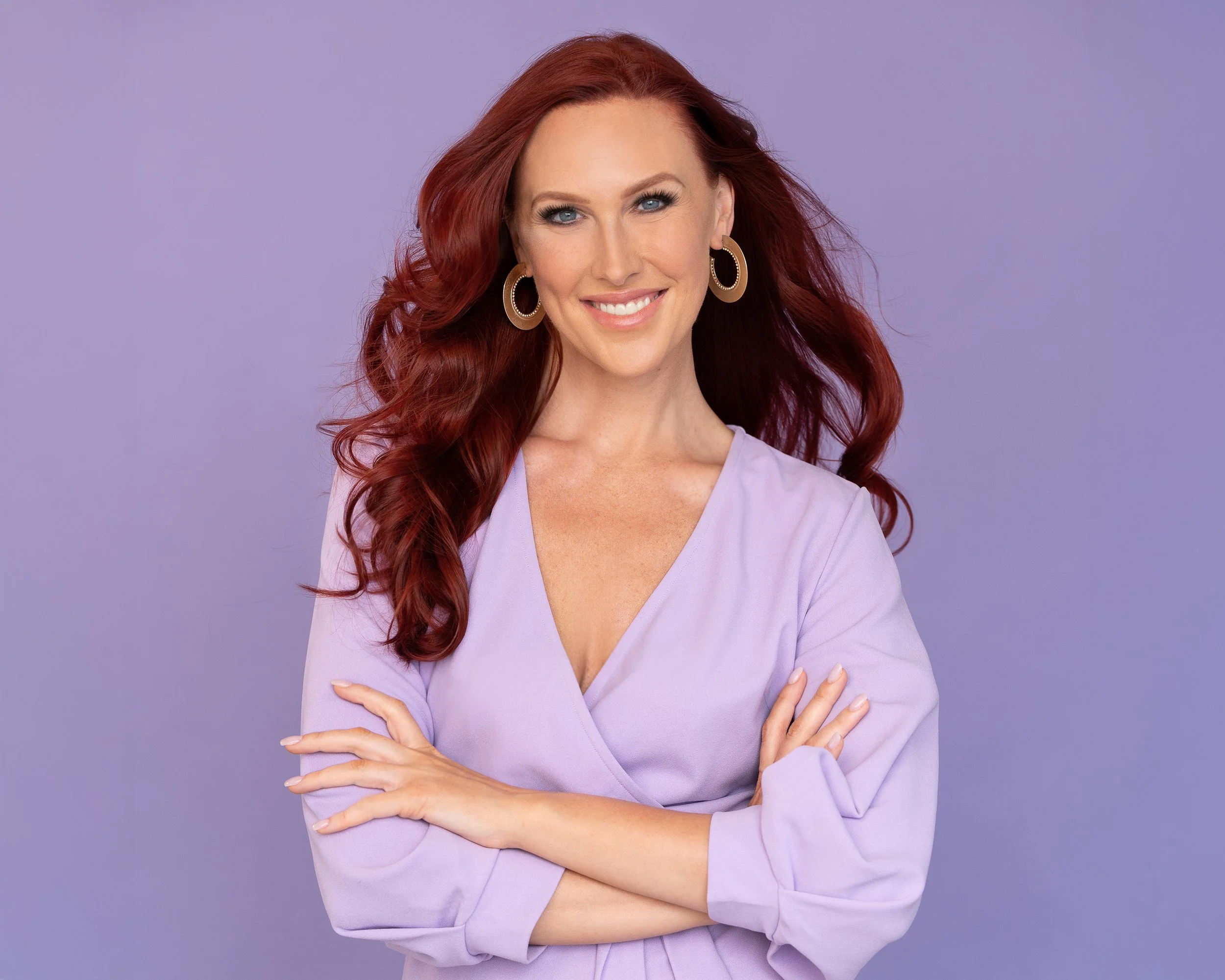 A woman with long red hair, blue eyes, and large gold and black earrings, smiling and crossing her arms, wearing a light purple top, against a purple background.