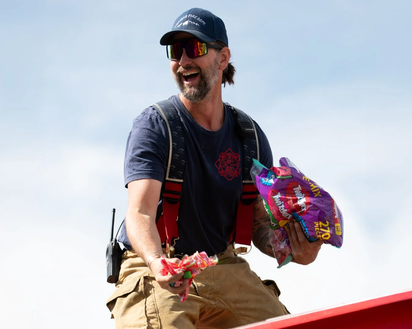 A firefighter smiling and holding a bag of snacks, wearing sunglasses, a cap, and a backpack, standing outdoors against a clear sky.