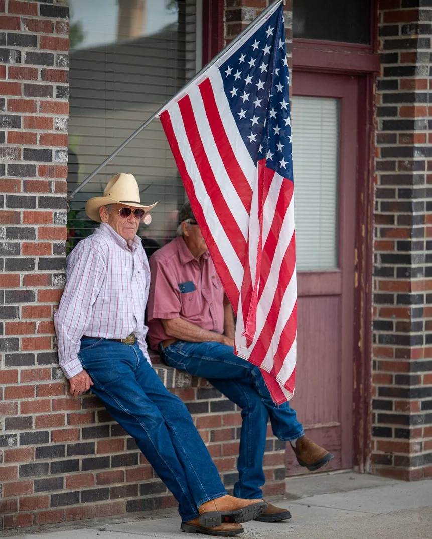Two elderly men sitting on a brick wall, one wearing a cowboy hat and sunglasses, and the other wearing glasses and a pink shirt, with an American flag hanging in front of them.