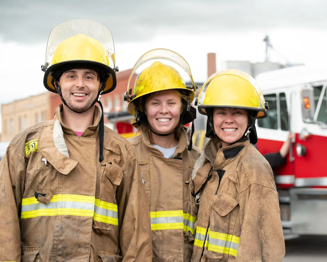 Three firefighters, two women and one man, wearing yellow helmets and brown firefighting suits, standing in front of a fire truck, smiling at the camera.