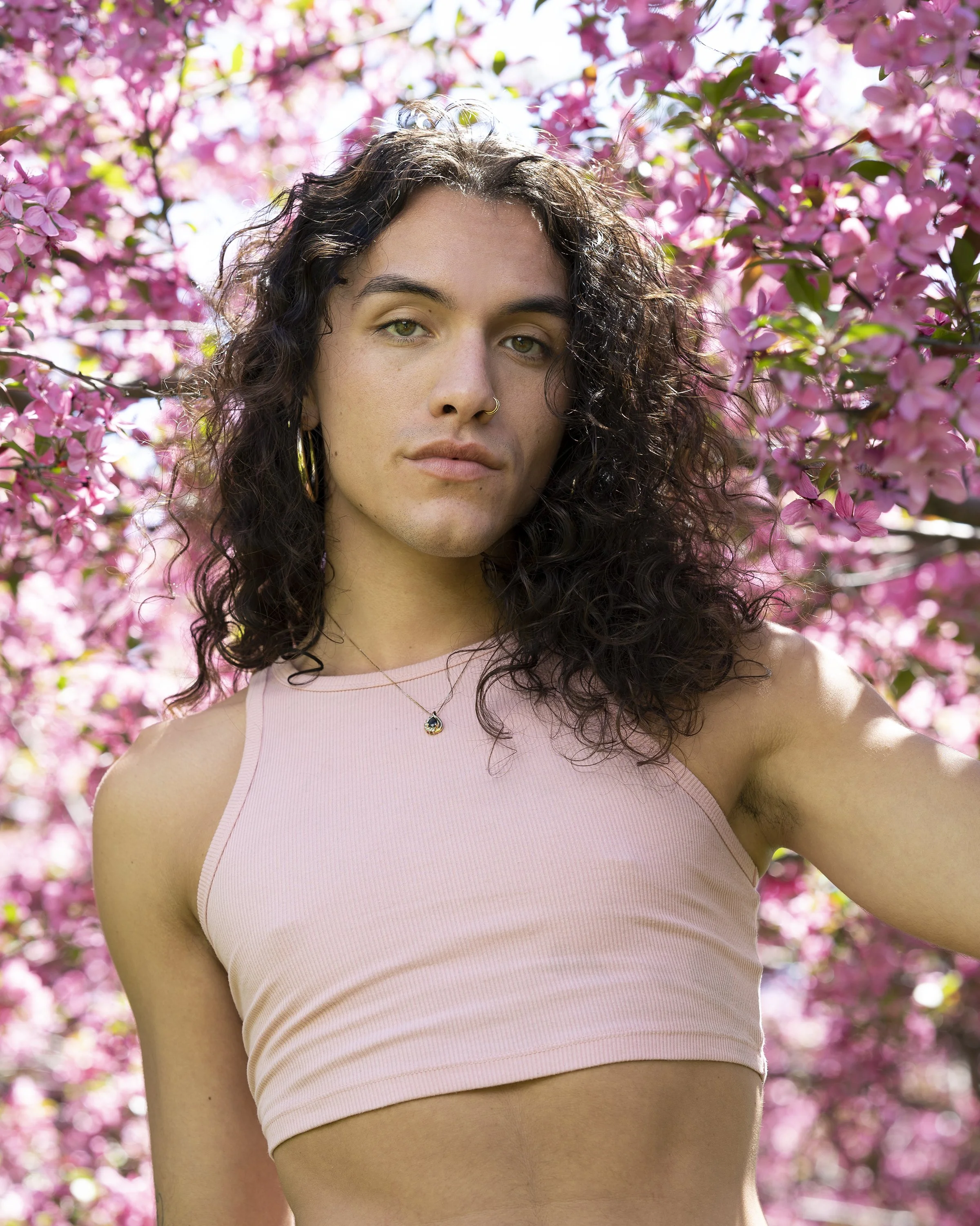 A young woman with curly dark hair, wearing a pink sleeveless top and a necklace with a small pendant, standing outdoors among pink flowering trees with sunlight filtering through the branches.