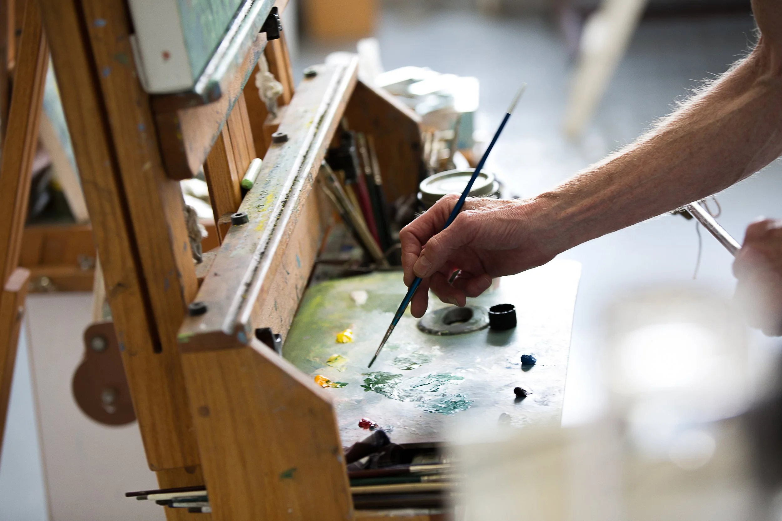 Close-up of an artist's hand holding a paintbrush, working on a colorful painting on a wooden easel in a studio.