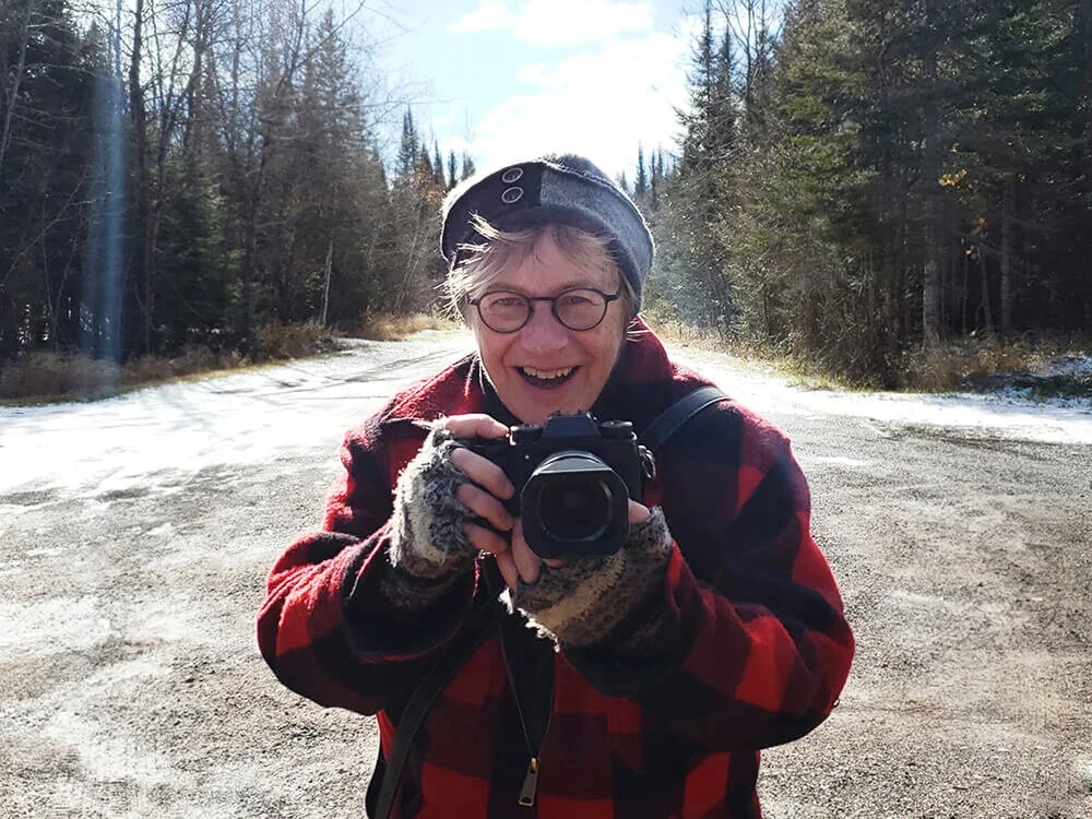 An elderly woman with glasses and short hair, wearing a beanie, red and black checkered jacket, and gloves, holding a camera with a wide smile outdoors on a gravel path surrounded by trees in a forest with snow on the ground.