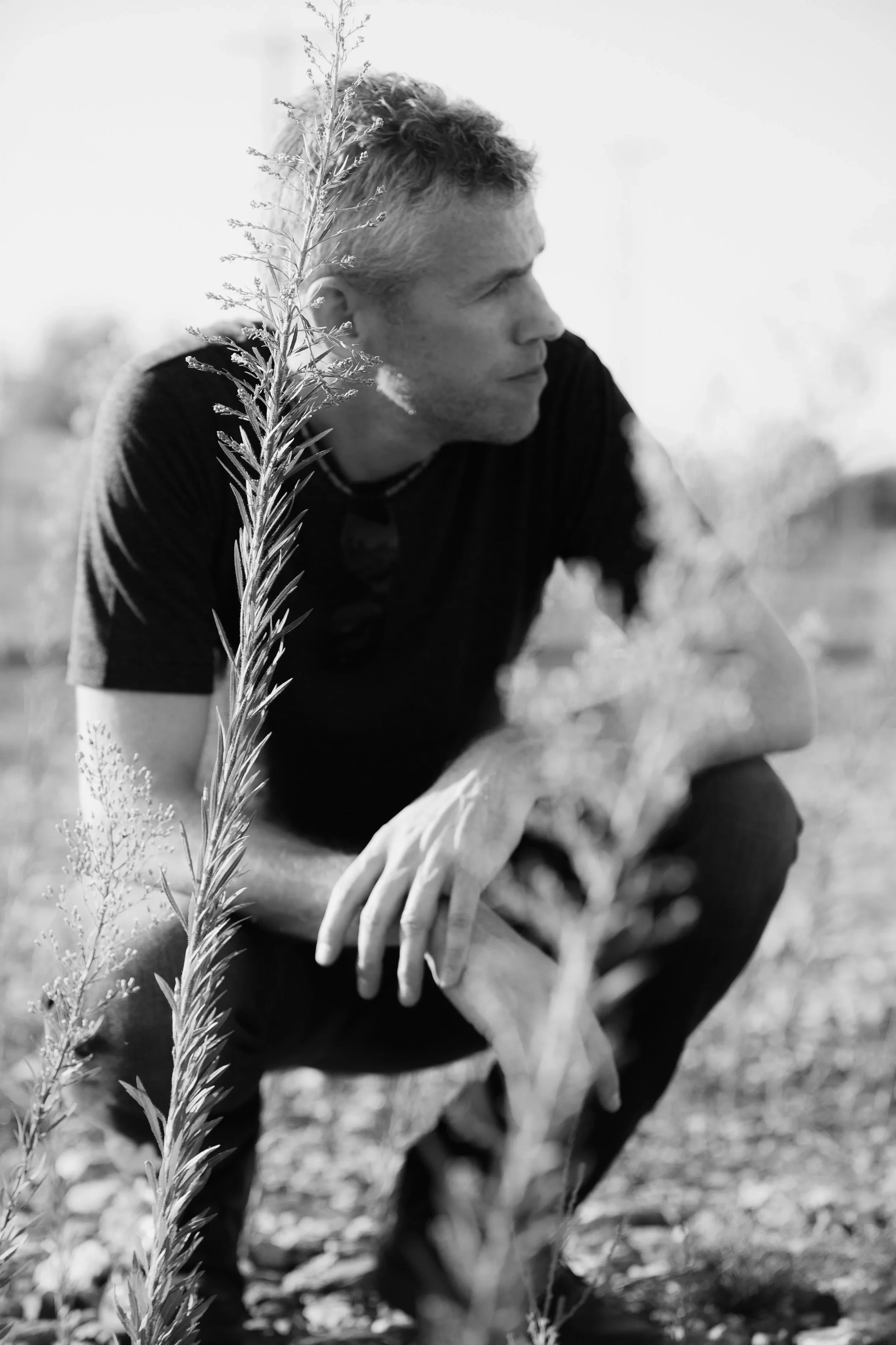 A black and white photo of a man crouching outdoors, wearing a dark T-shirt and a glove on his right hand, with a tall plant in the foreground.