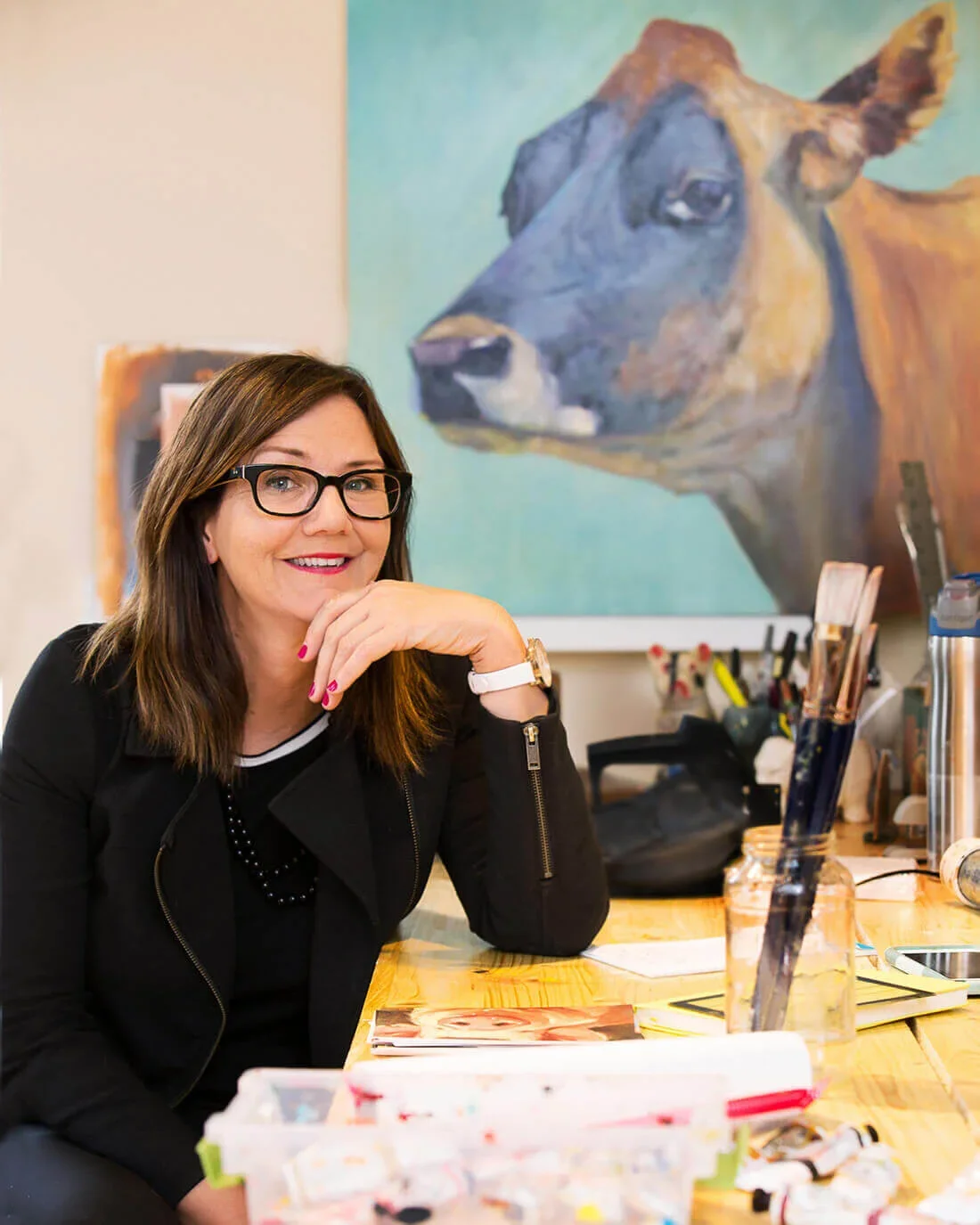 A woman with glasses and brown hair smiling and resting her chin on her hand in an art studio with a large cow painting behind her.