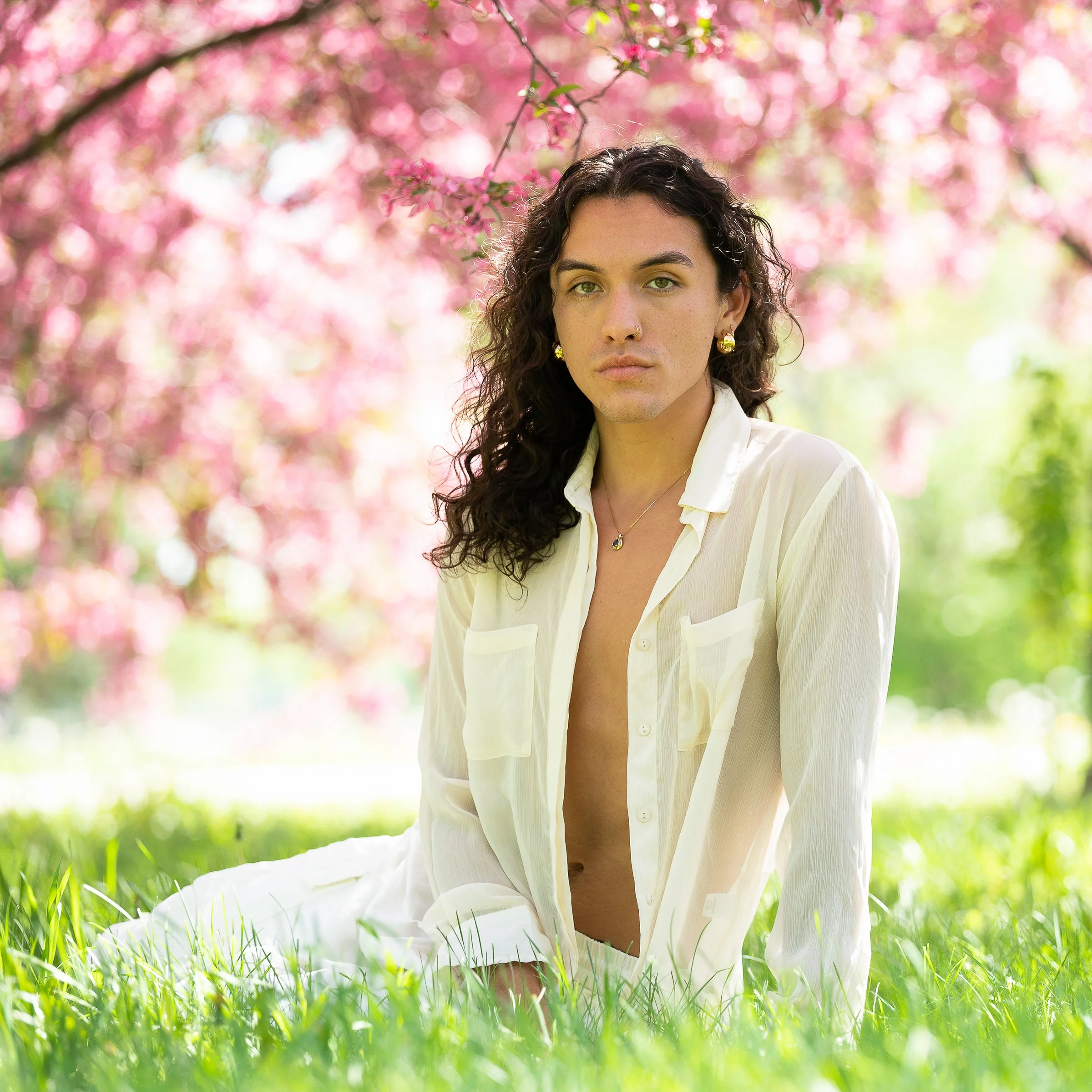 A woman with curly dark hair, wearing a white shirt, sitting on green grass in a park with pink blooming trees in the background.