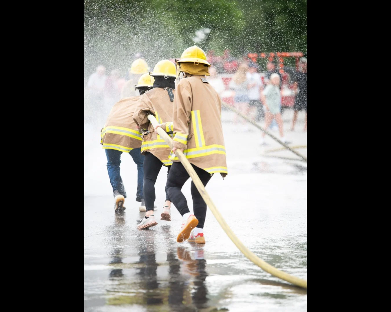 Group of firefighters in yellow helmets and brown protective gear battling a fire with a hose, while spectators watch behind a safety barrier.