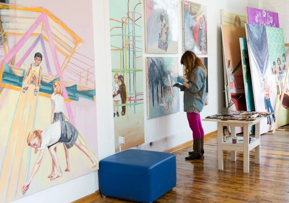 An artist painting colorful portraits and scenes in a gallery space, with additional paintings on display and art supplies on a small table.