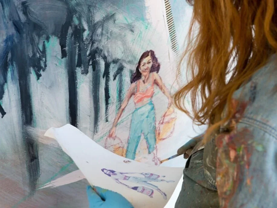 A person with red hair painting a mural of a woman with dark hair wearing an orange top and blue jeans, standing in a park-like setting with trees.