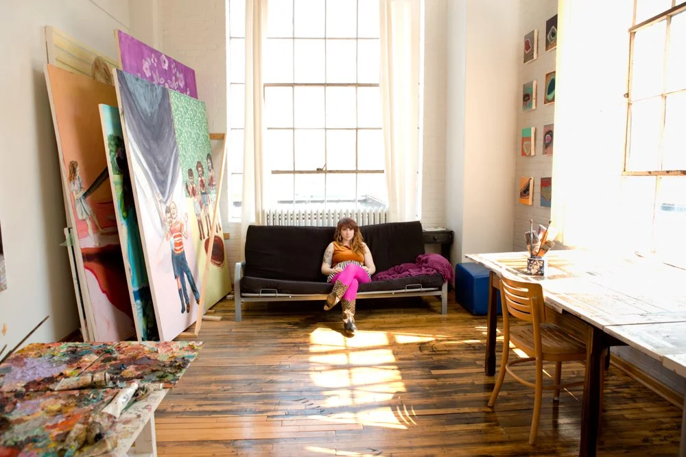 An artist's studio with large colorful paintings leaning against the wall, a woman sitting on a black sofa, and a wooden table with art supplies near a large window.