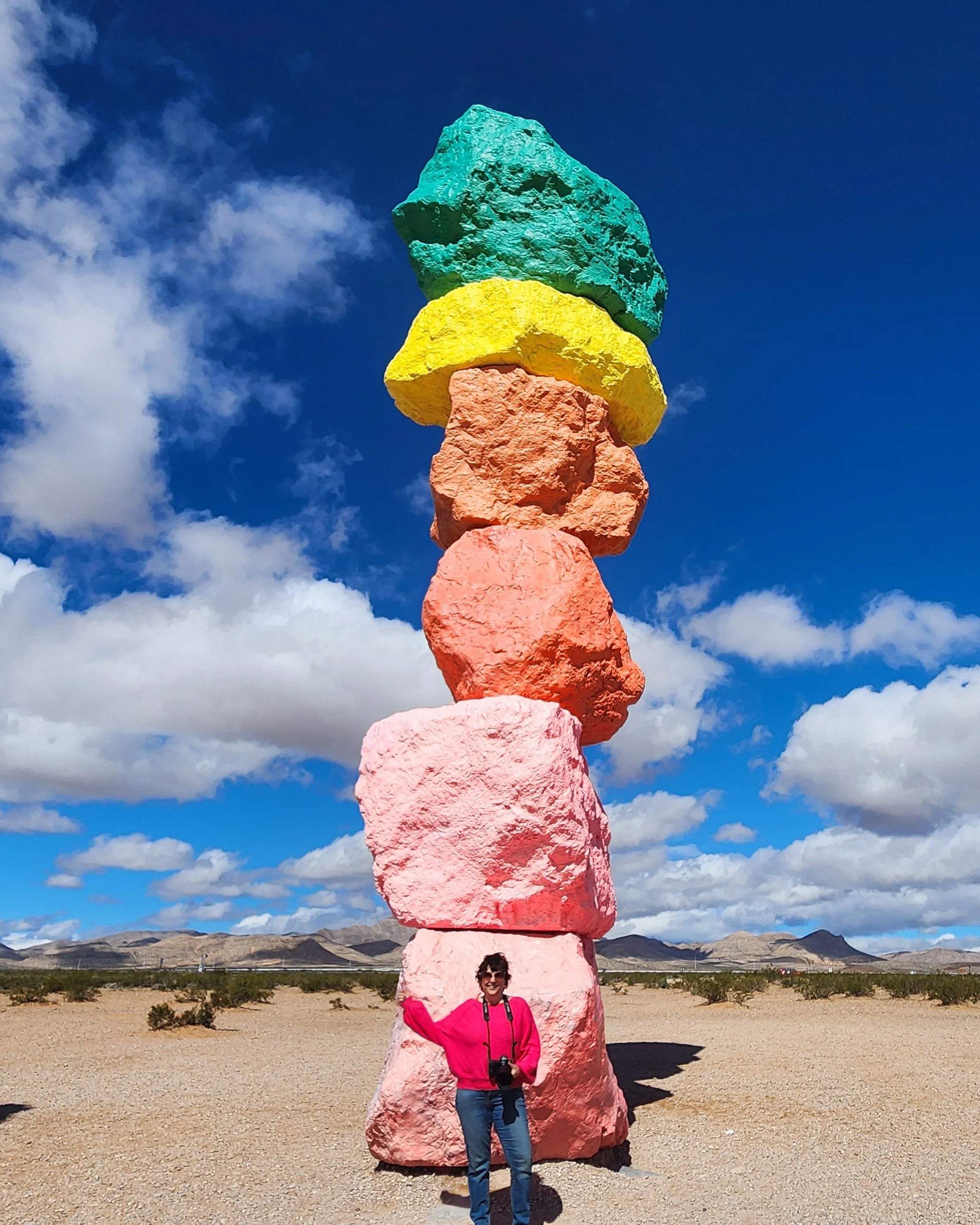A woman in a pink jacket and blue jeans standing in front of a large, colorful rock formation in a desert landscape with mountains on the horizon under a partly cloudy blue sky.