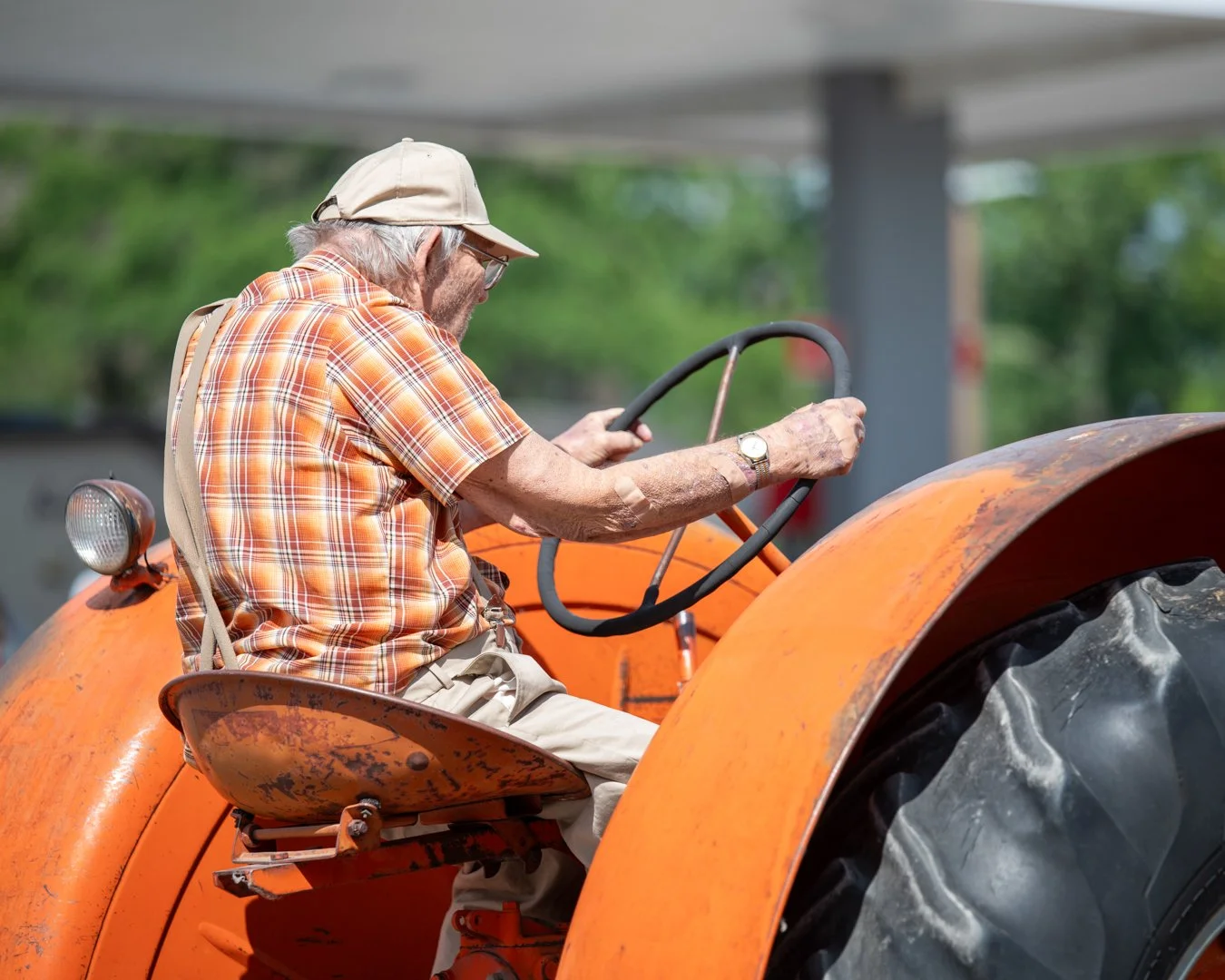 An elderly man with gray hair, glasses, and a beige cap driving an orange tractor wearing a plaid shirt and beige pants.