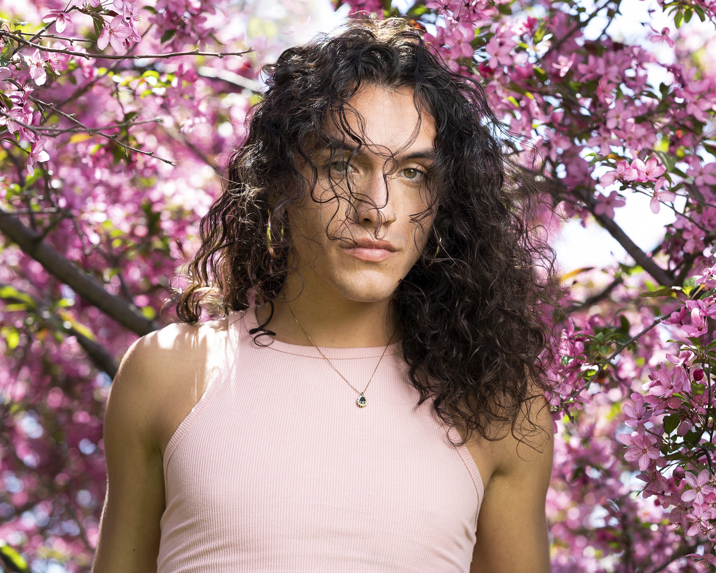 A woman with curly dark hair and light skin, wearing a pink sleeveless top and a necklace, standing outdoors in front of a blooming pink flowering tree.