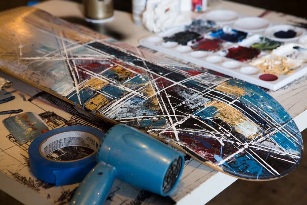 An artist's workspace with a skateboard painted in an abstract style, a blue hairdryer, masking tape, and a paint palette with various colors.