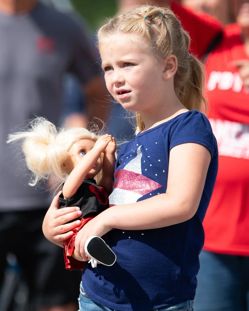 A young girl holding a blonde doll with a ponytail, outside on a sunny day, wearing a navy blue t-shirt with a star and black shoes.