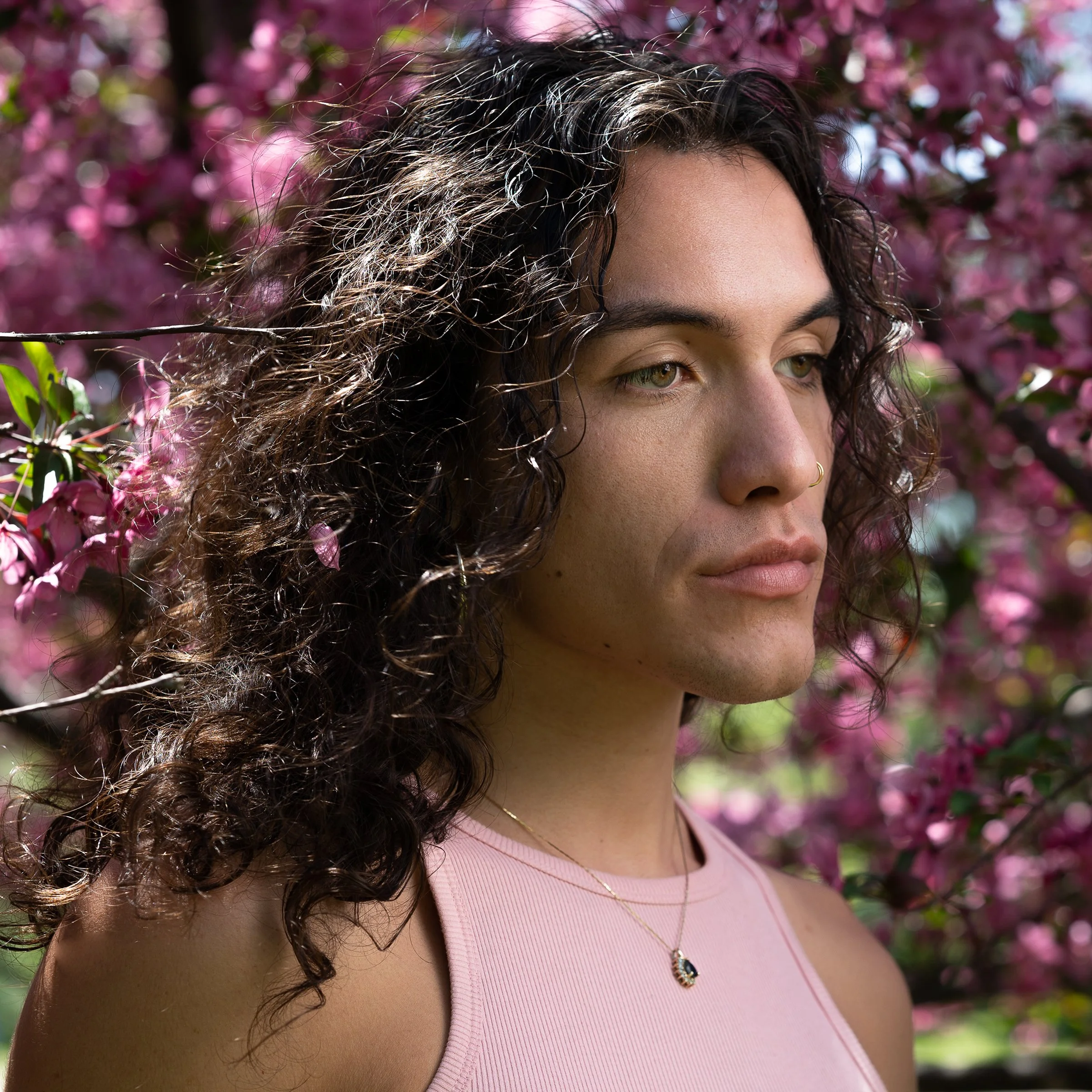 Close-up of a woman with curly dark hair and eyes, wearing a pink top and a gold necklace with a pendant, standing outdoors with pink flowering bushes in the background.