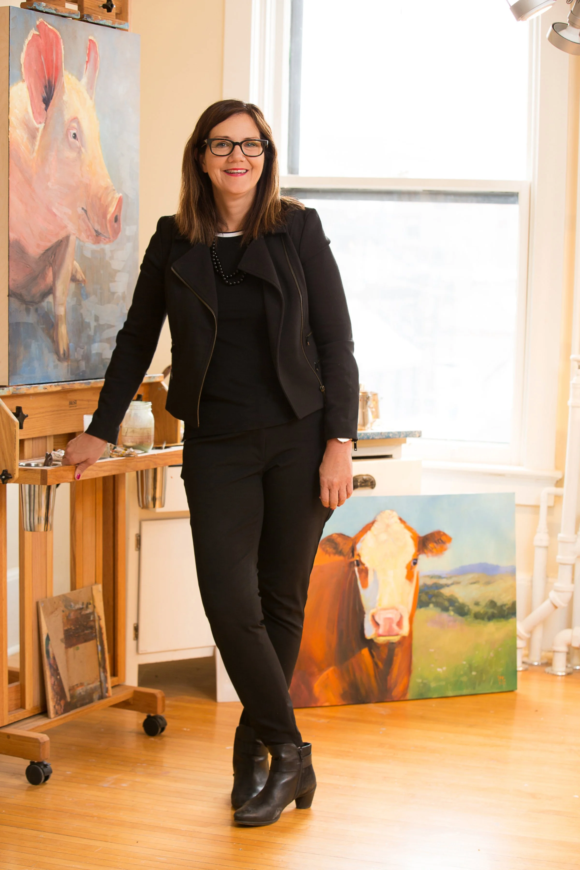 A woman standing in an art studio, smiling, wearing black glasses, a black jacket, and black pants, with a cowboy boot on one foot. Behind her are paintings of a pig and a cow, along with art supplies and a window.