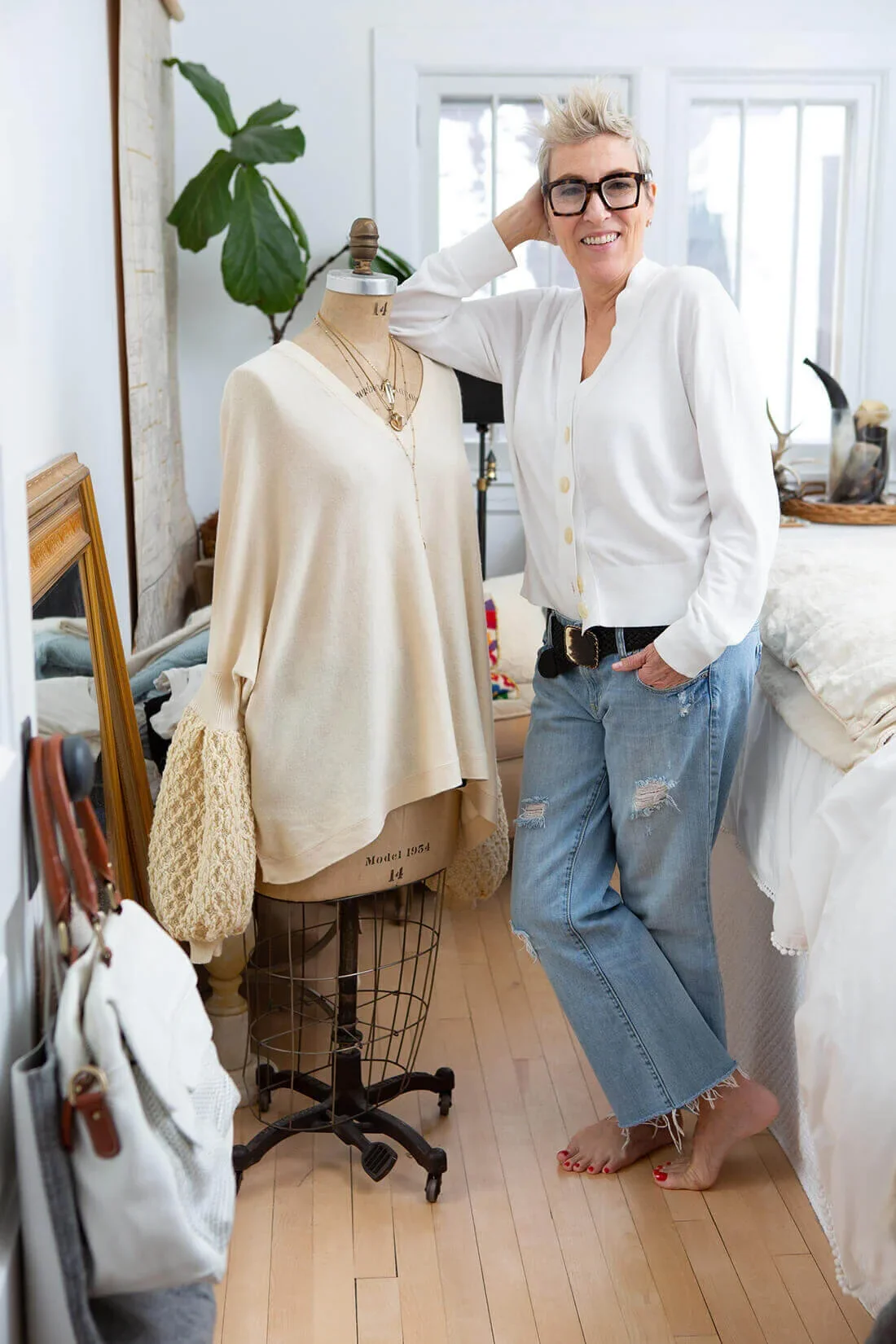 A woman with short blonde hair, glasses, and a white button-up shirt stands barefoot next to a dress form mannequin displaying layered necklaces and a cream-colored sweater. She is smiling and has her hand resting on her head in a cozy, well-lit room