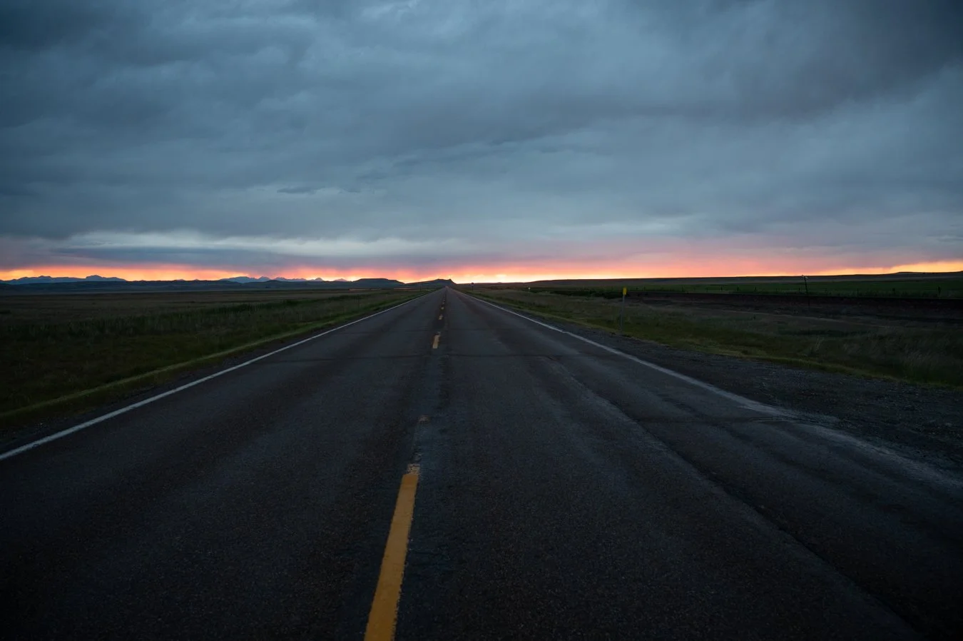 Open road stretching into the distance under a cloudy sky at sunset, with mountains on the horizon.