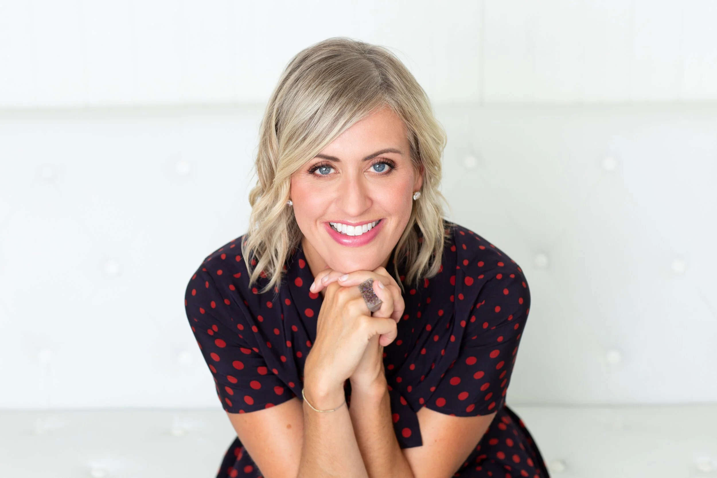 A woman with blonde hair and blue eyes, smiling, wearing a dark dress with red polka dots, sitting with her chin resting on her hands.