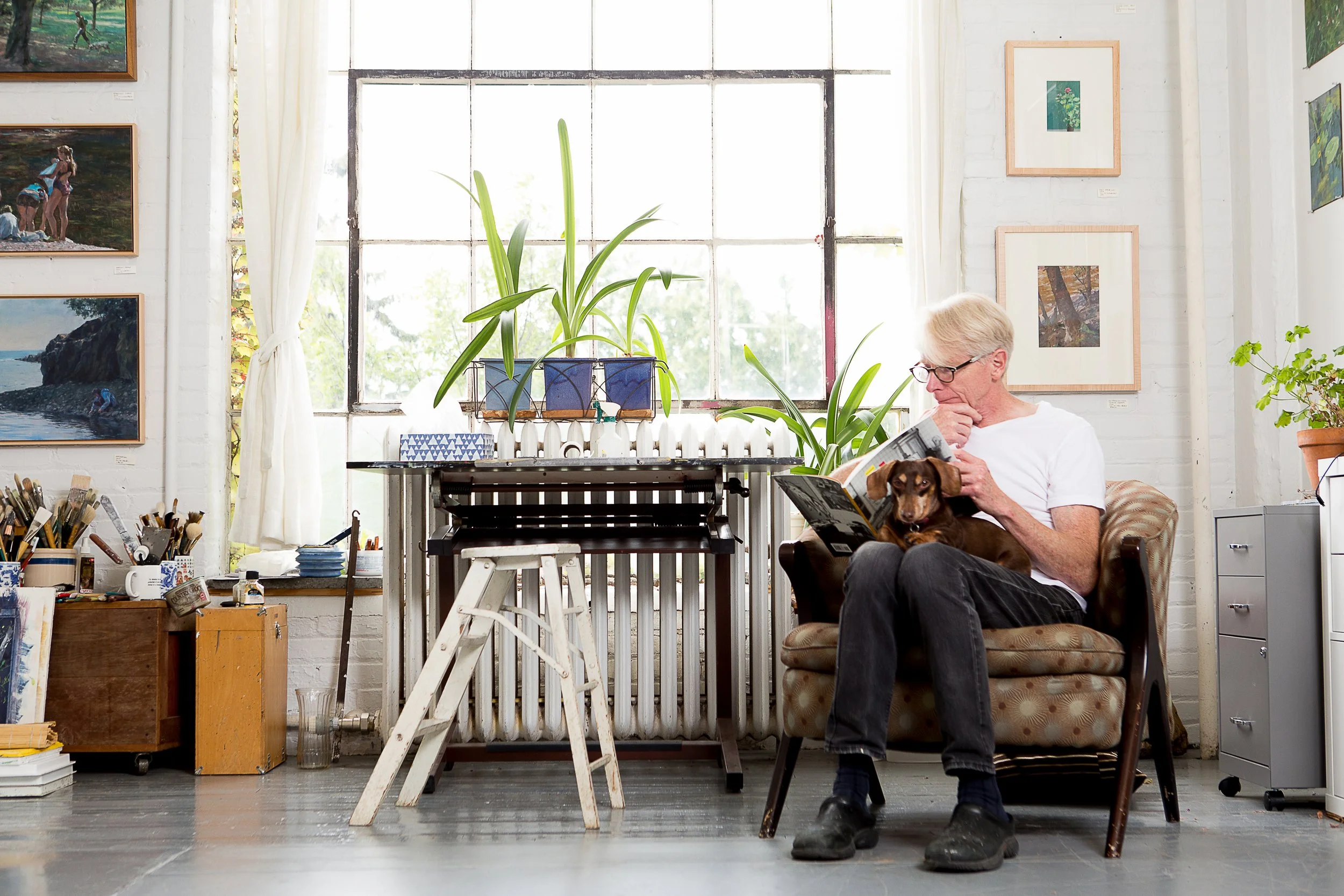 An elderly man with gray hair and glasses reading a magazine while sitting on a patterned armchair, with a brown dog on his lap, in an art studio with paintings on white brick walls, potted plants, and art supplies.