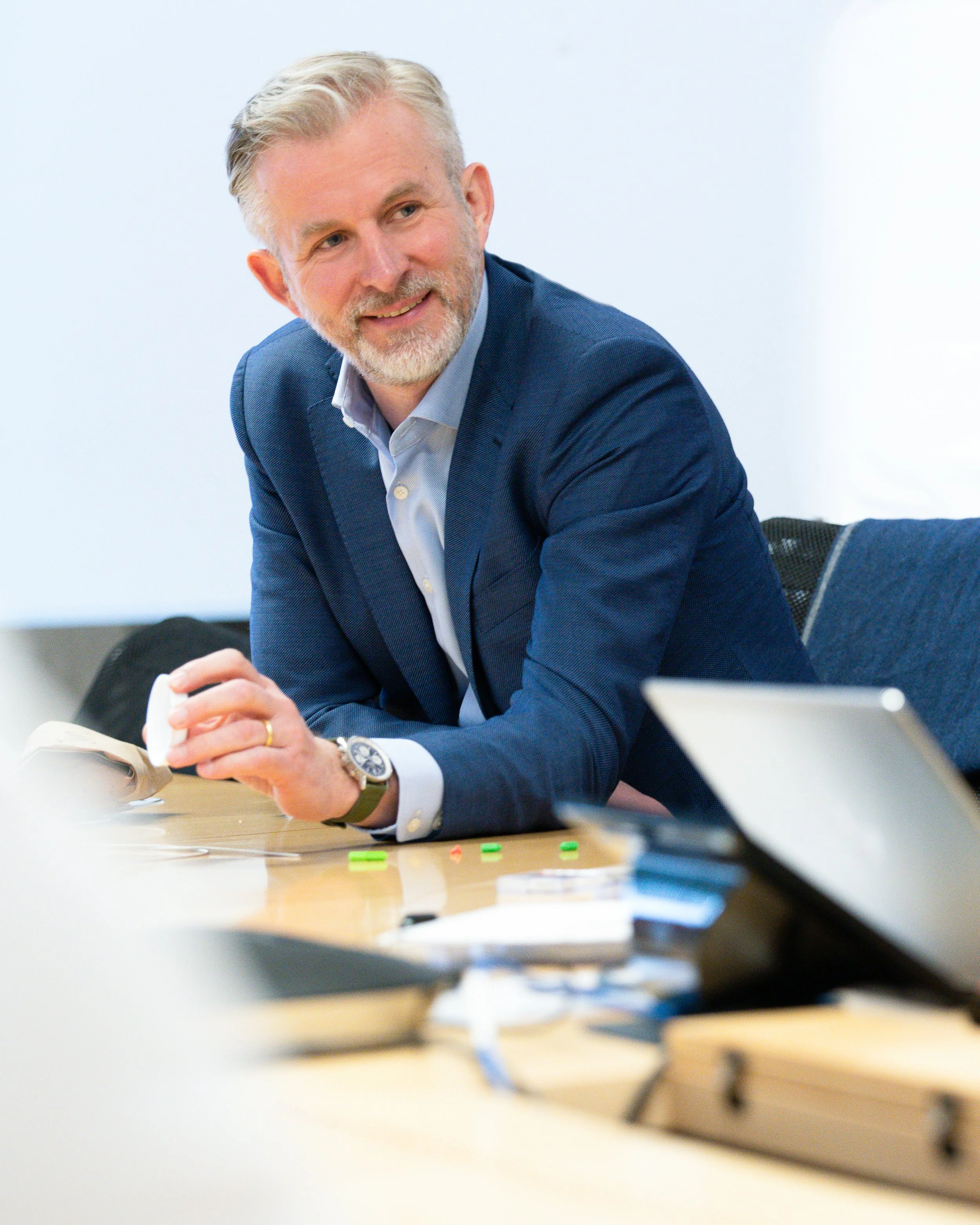 A middle-aged man with gray hair and beard, wearing a blue suit and watch, smiling and sitting at a table in a professional setting, with papers and a laptop in front.