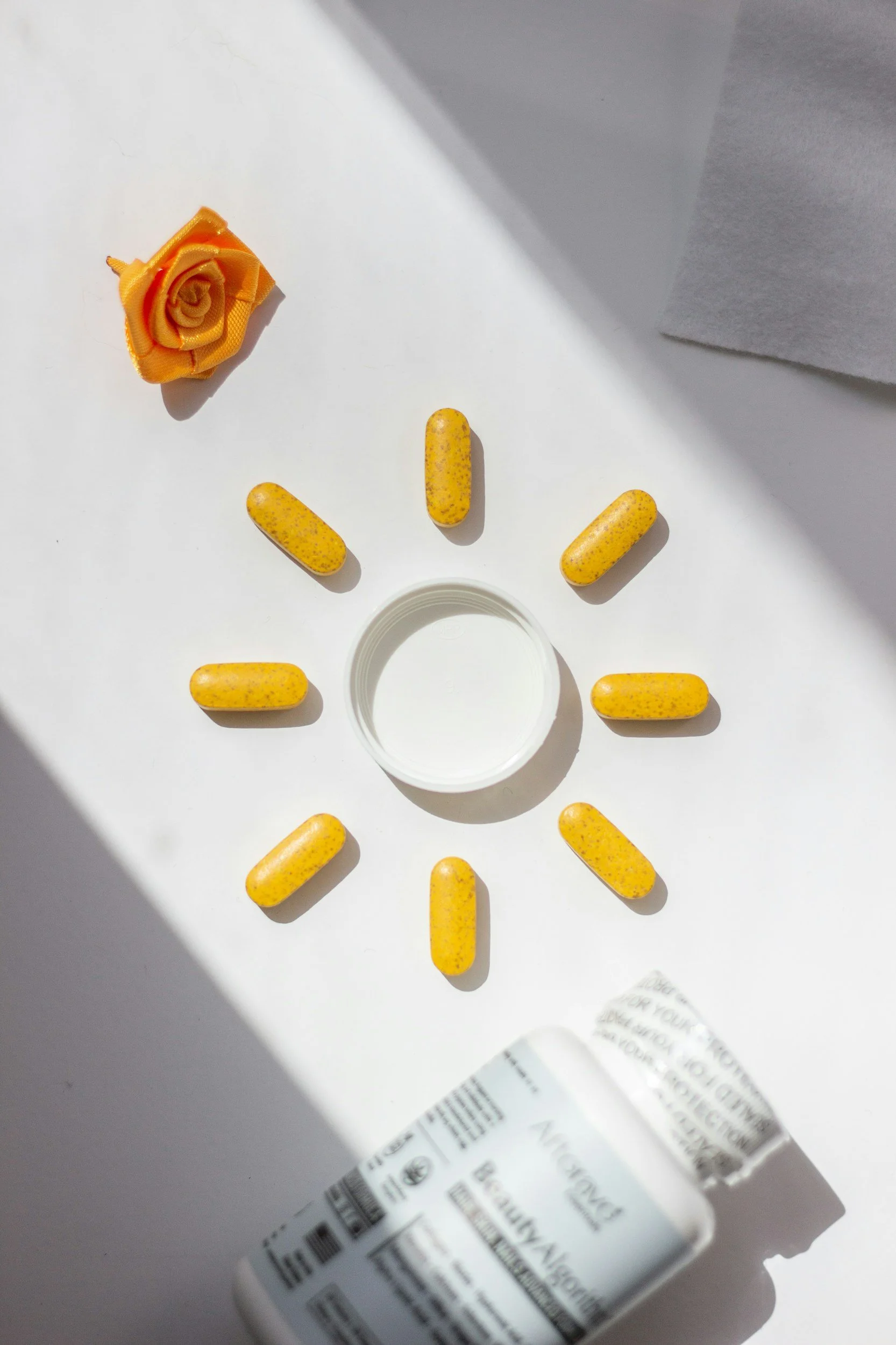 Yellow speckled capsules arranged in a semi-circle on a white surface with an orange fabric rose and a white bottle cap, all in natural sunlight.