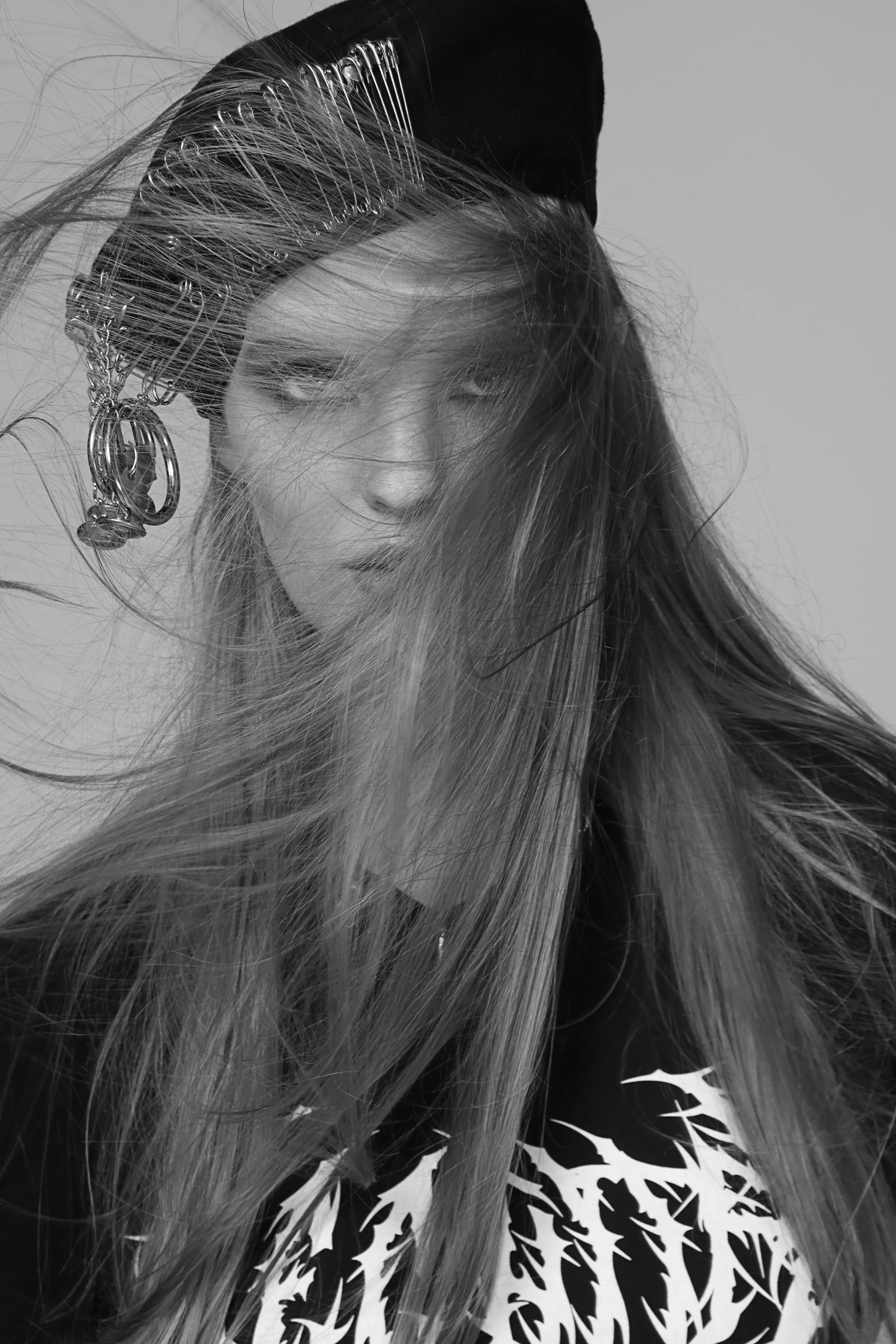 Black and white photo of a person with long hair covering part of their face, wearing a hat with safety pins and a patterned shirt.
