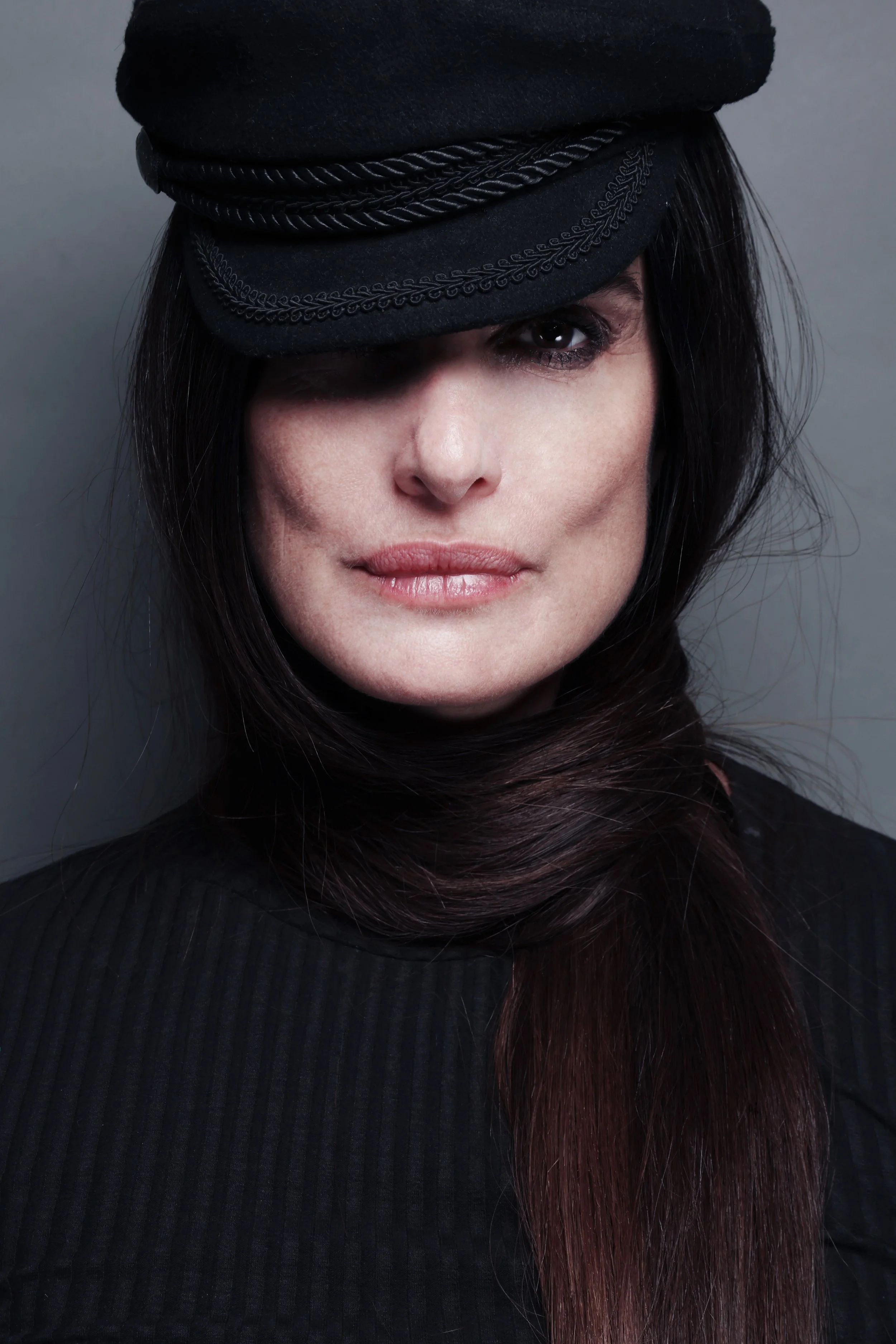 A woman with dark hair, wearing a black cap with decorative stitching, a braided band, and a black turtleneck.