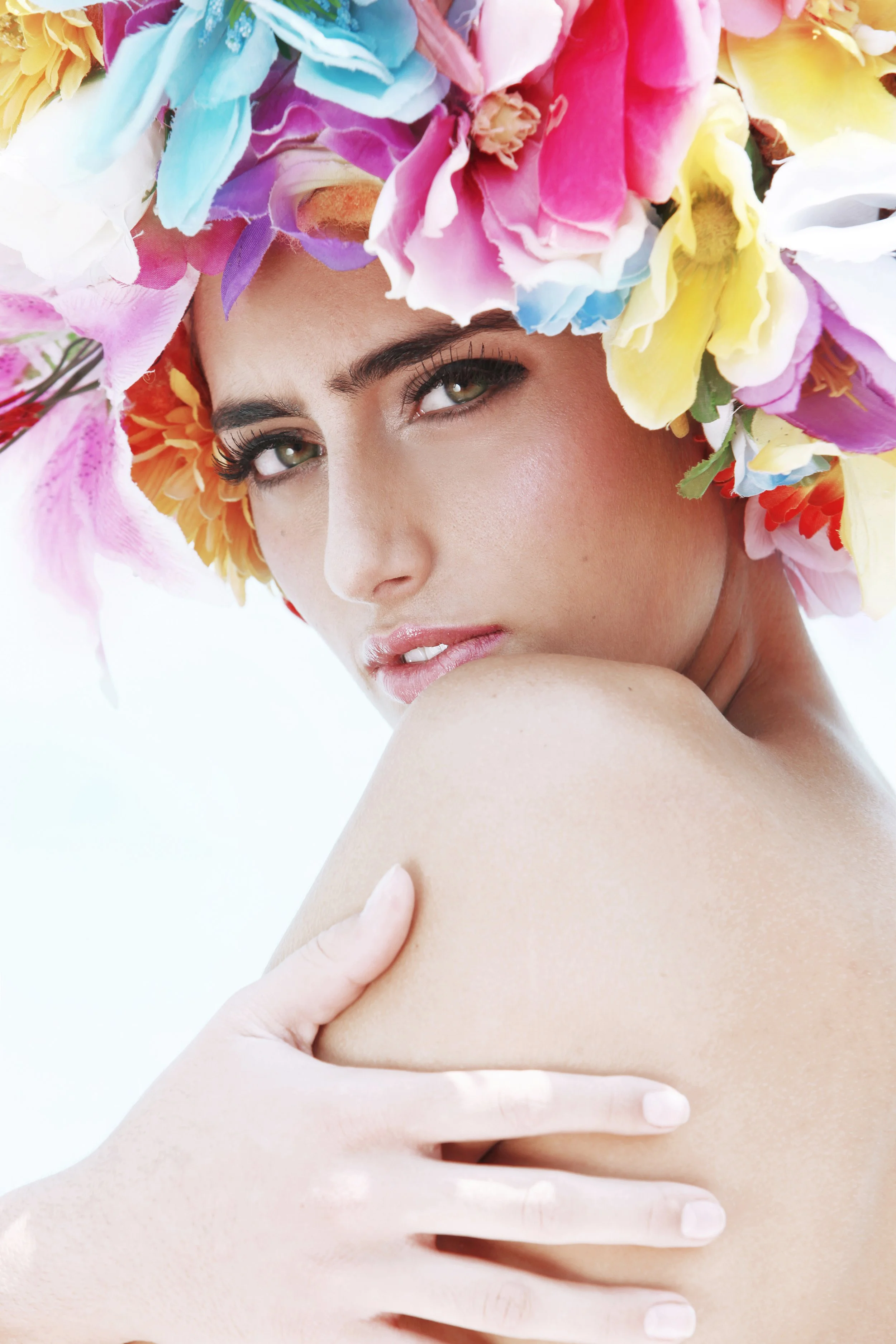 Close-up photo of a woman with a colorful flower crown made of pink, yellow, blue, purple, and orange flowers. She has fair skin, green eyes, long eyelashes, and light makeup, looking directly at the camera with a soft expression. Her hand is resting