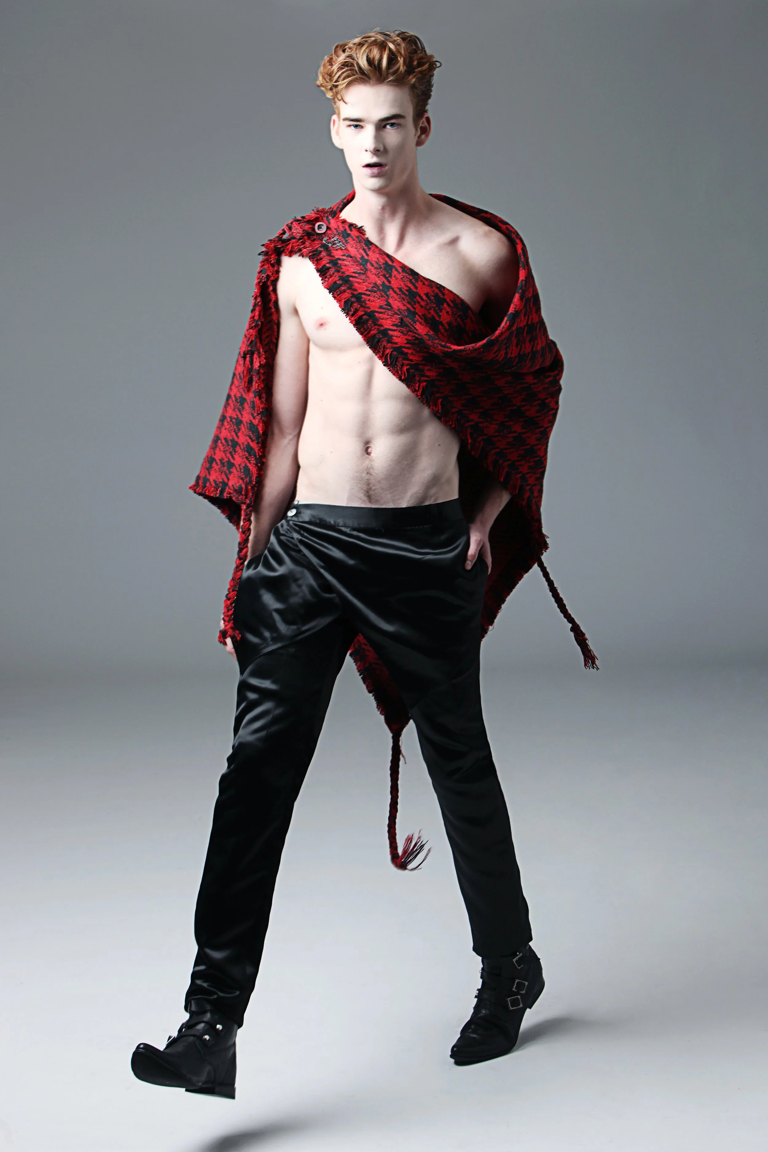 Young man with curly red hair wearing black boots, black pants, and a red and black patterned shawl draped over one shoulder while standing against a plain gray background.