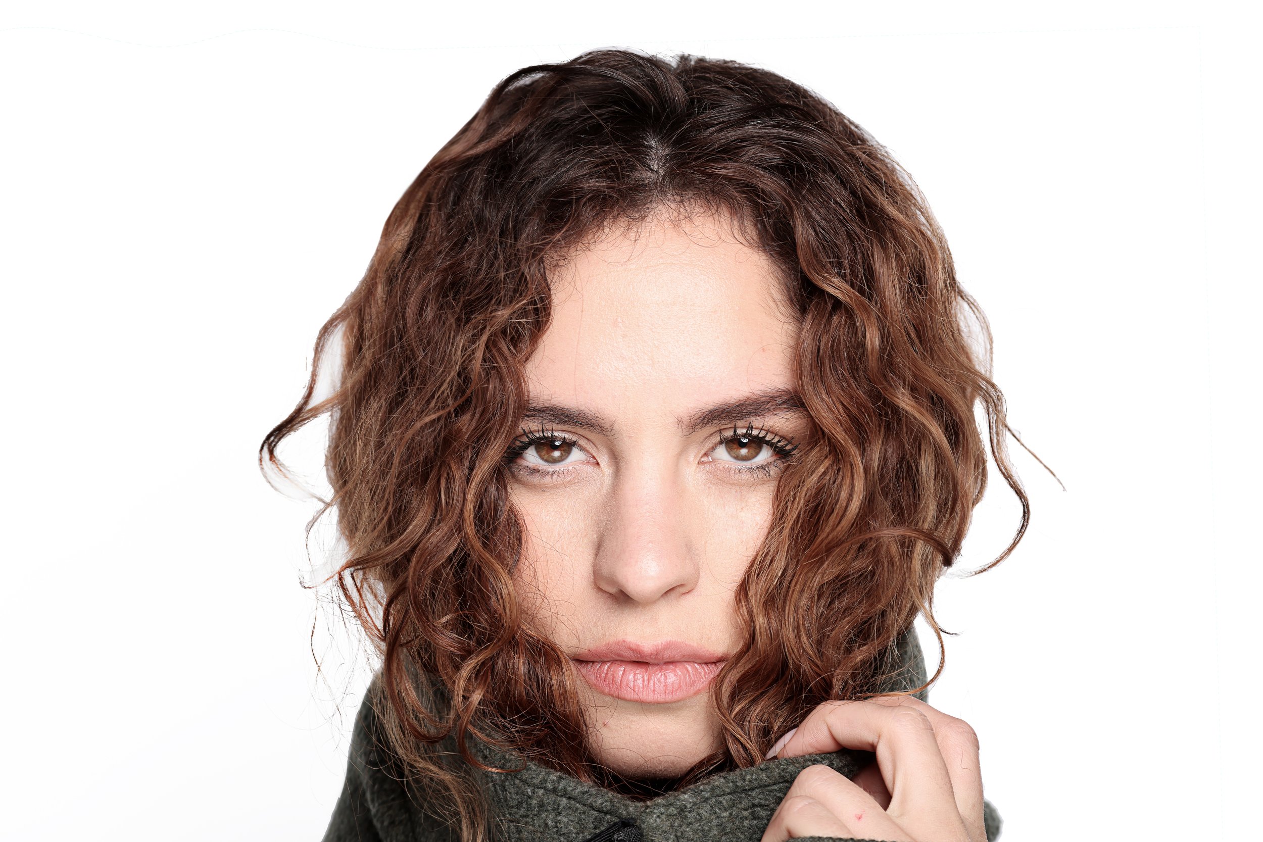 Close-up of a woman with curly brown hair, visible brown eyes, and light skin, wearing a dark gray sweater, looking at the camera, resting her chin on her hand.