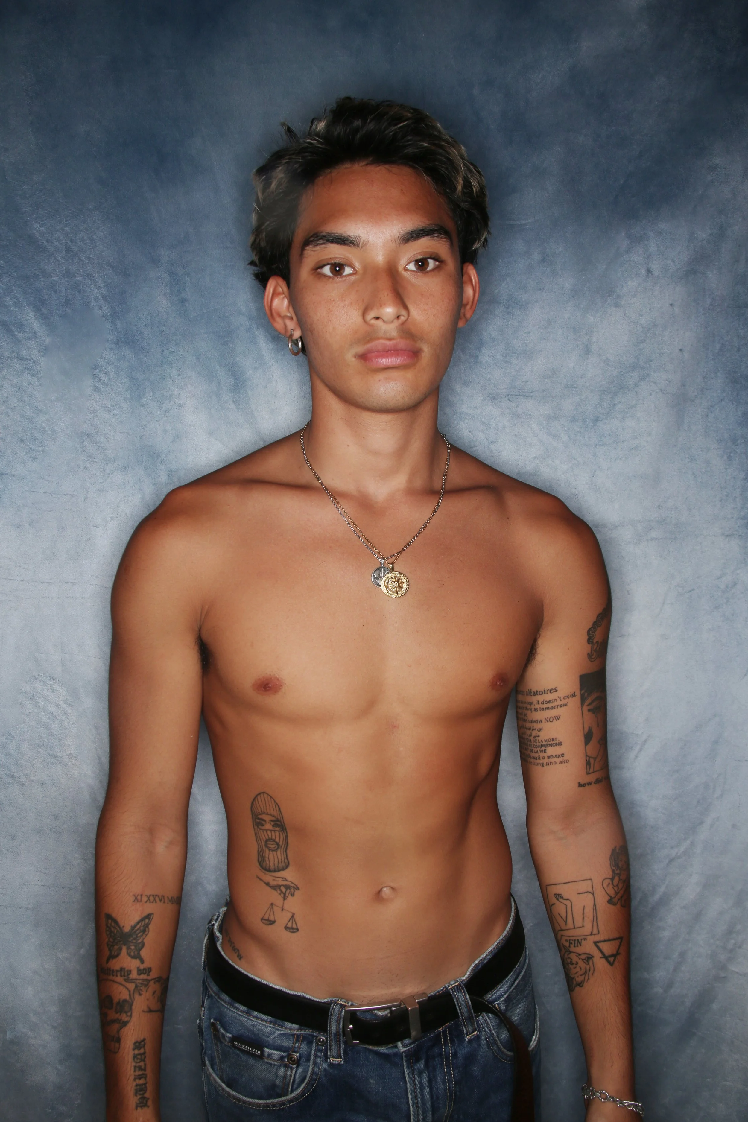 A shirtless young man with dark, styled hair and brown eyes, wearing multiple necklaces, earrings, and bracelets, has tattoos on his arms, torso, and ribs, and is standing against a plain, gray background with a serious expression.