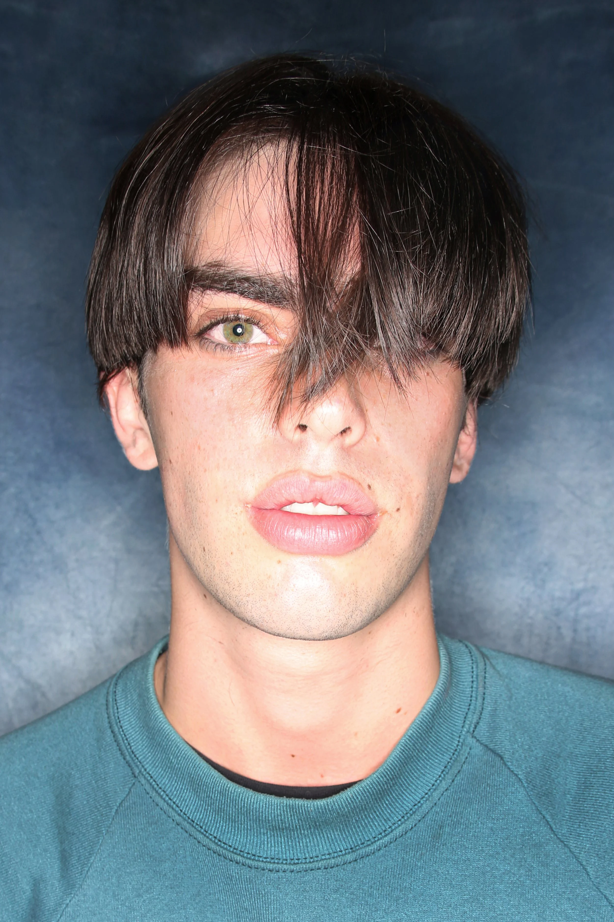 A young man with uneven dark brown hair partly covering one eye, green eyes, and light skin, wearing a teal shirt, against a plain background.