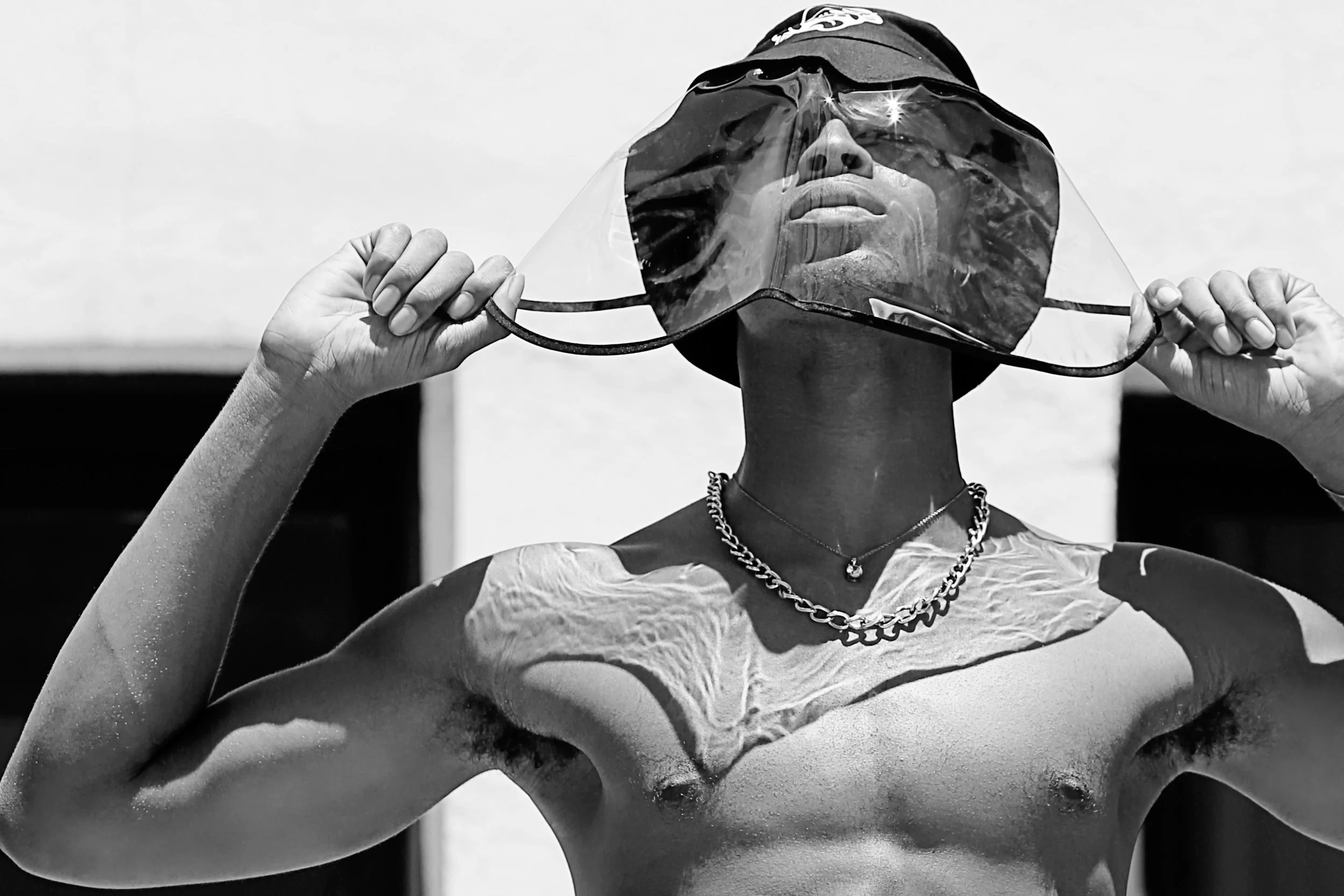 A person wearing sunglasses and a bandana, holding the sunglasses with both hands, taken from a slightly low angle in black and white.
