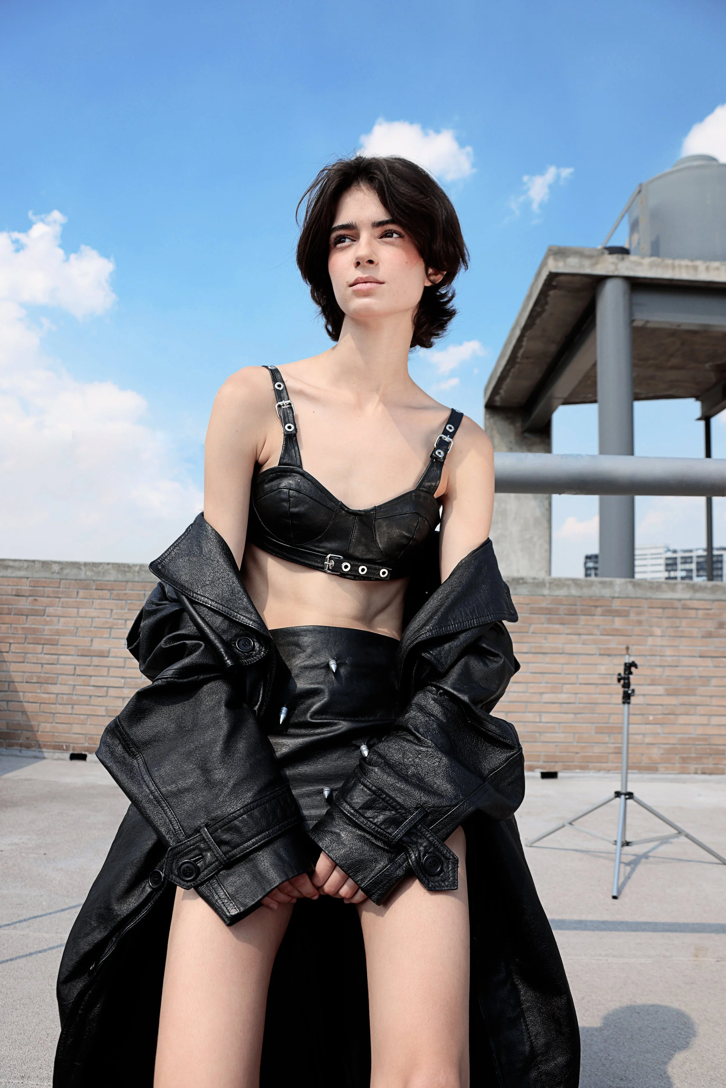 A young woman with short dark hair standing outdoors on a rooftop with a blue sky and some clouds. She is wearing a black leather bralette and black leather jacket, looking confidently into the distance.
