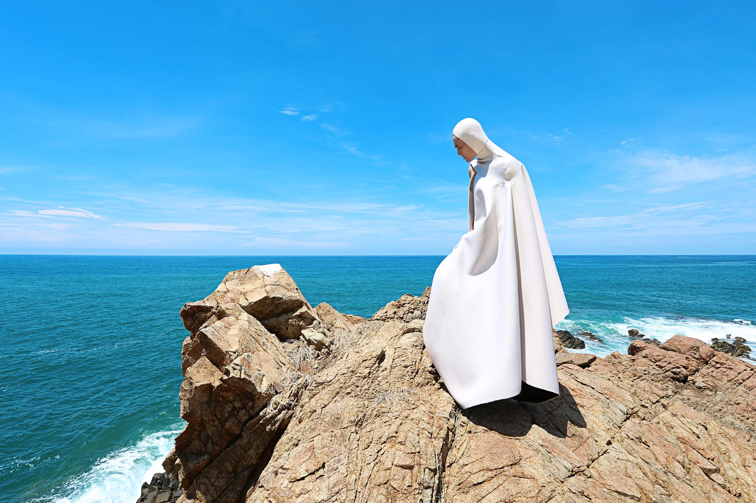 A person dressed in white standing on rocks by the ocean under a blue sky.