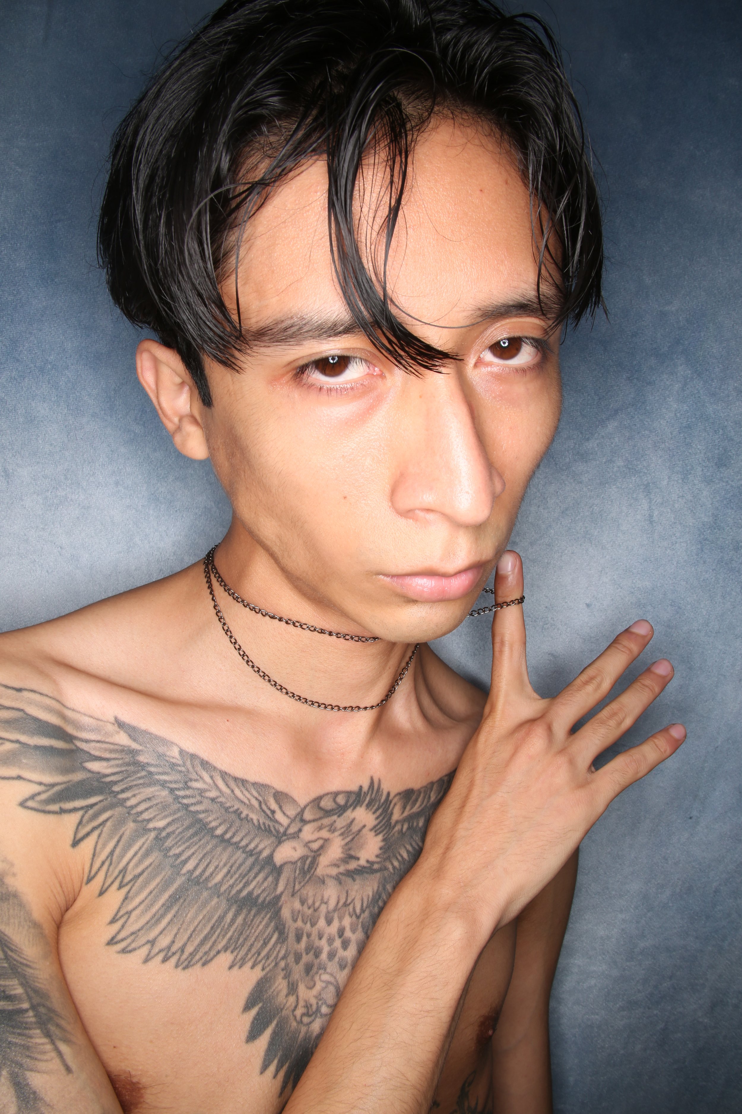 A shirtless man with black, wet hair, brown eyes, and a tattoo of an eagle on his chest, posing with his right hand near his face against a gray background.