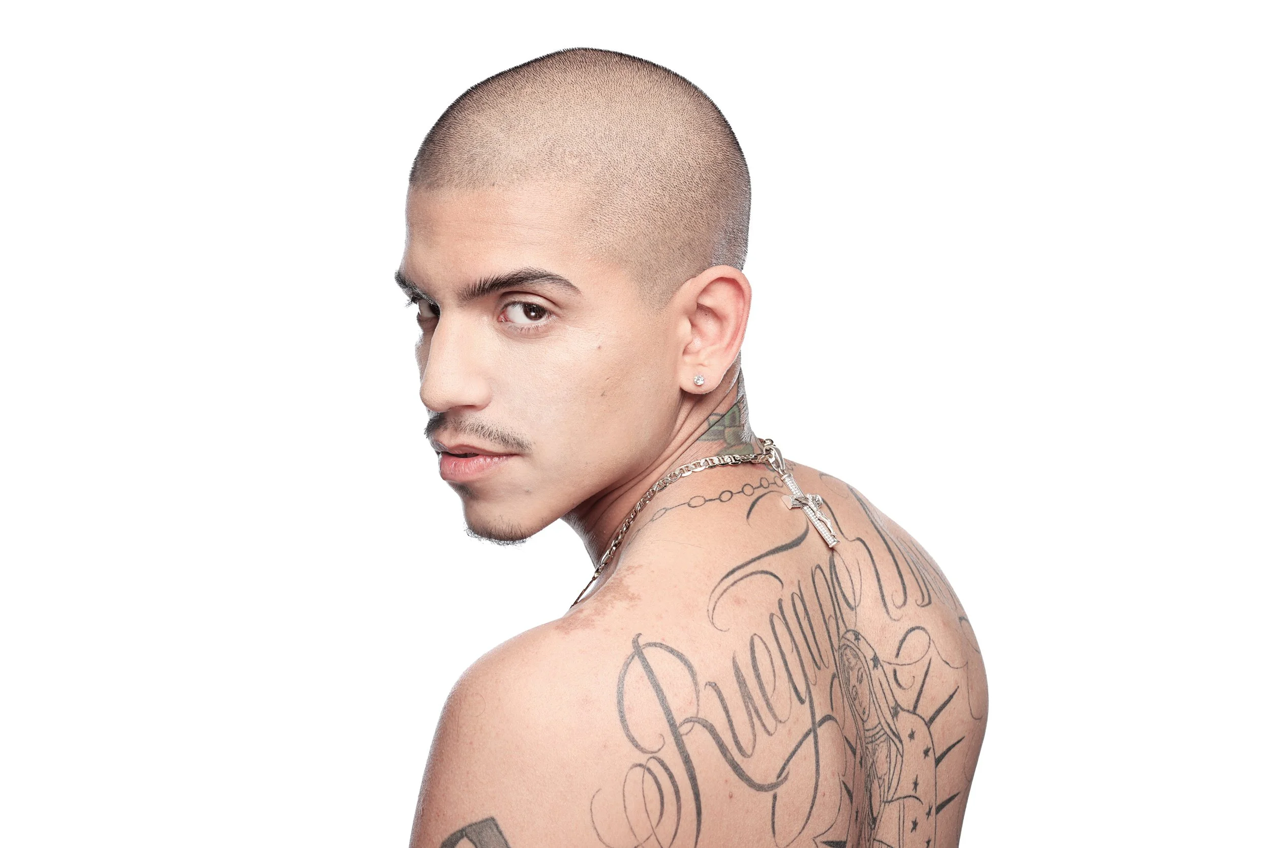Young man with a shaved head, tattoos on his neck and shoulder, wearing a diamond earring and a chain necklace, looking over his shoulder against a white background.