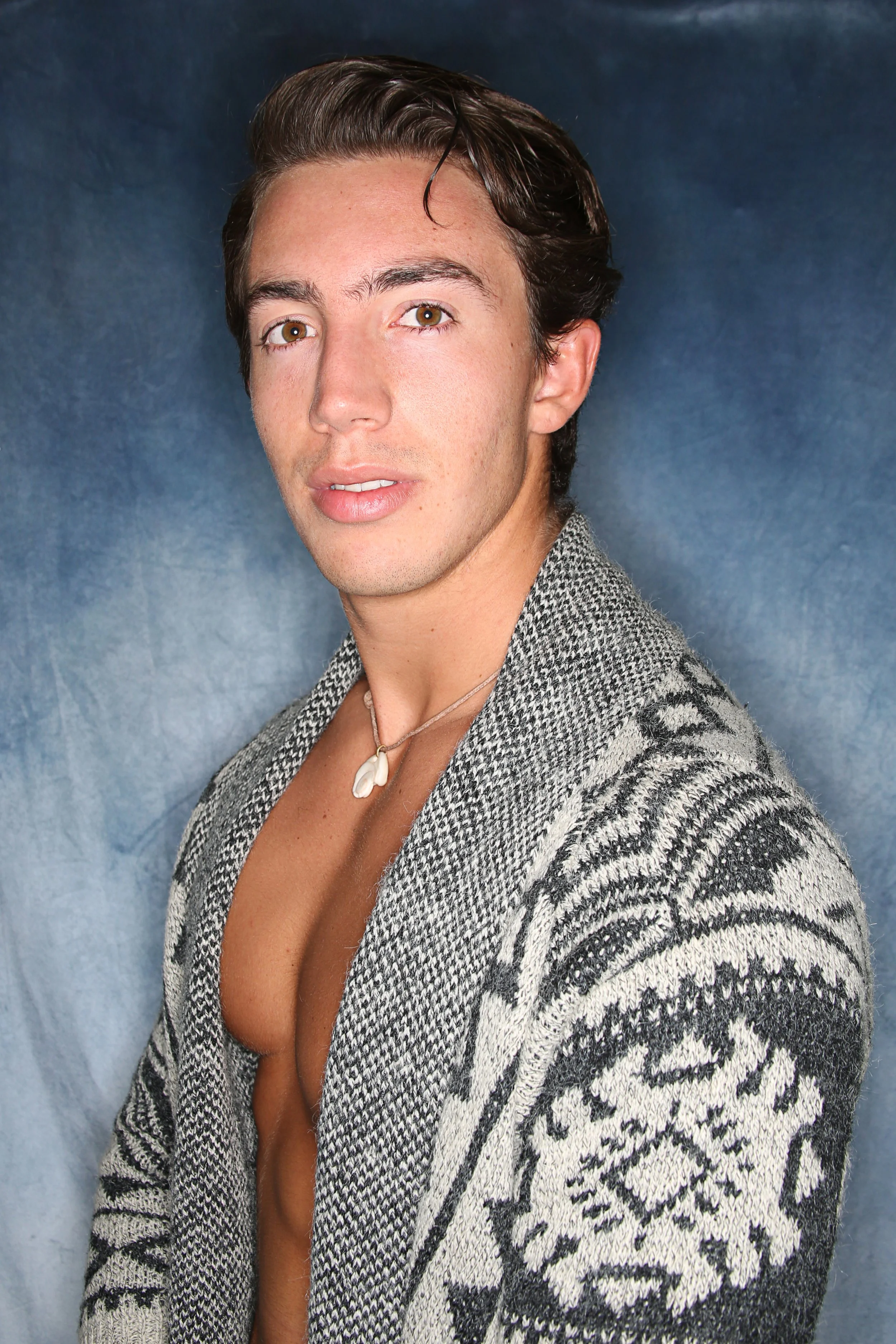 Close-up of a young man with brown hair and brown eyes wearing a patterned sweater and a shell necklace, standing against a blue background.