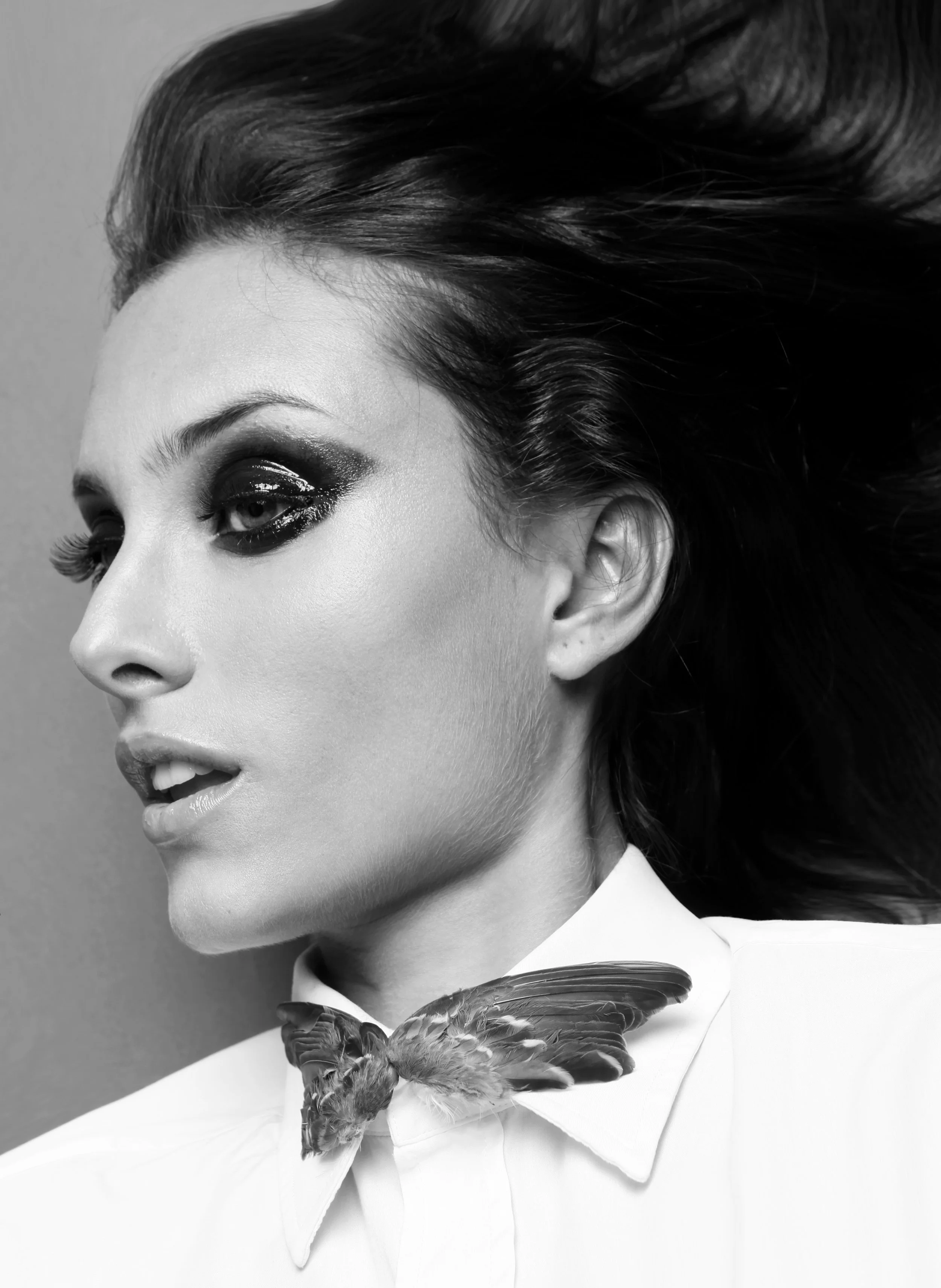 Black and white close-up of a woman's face, showing her eye makeup, lips, and cheek, with a bird perched on her collar.