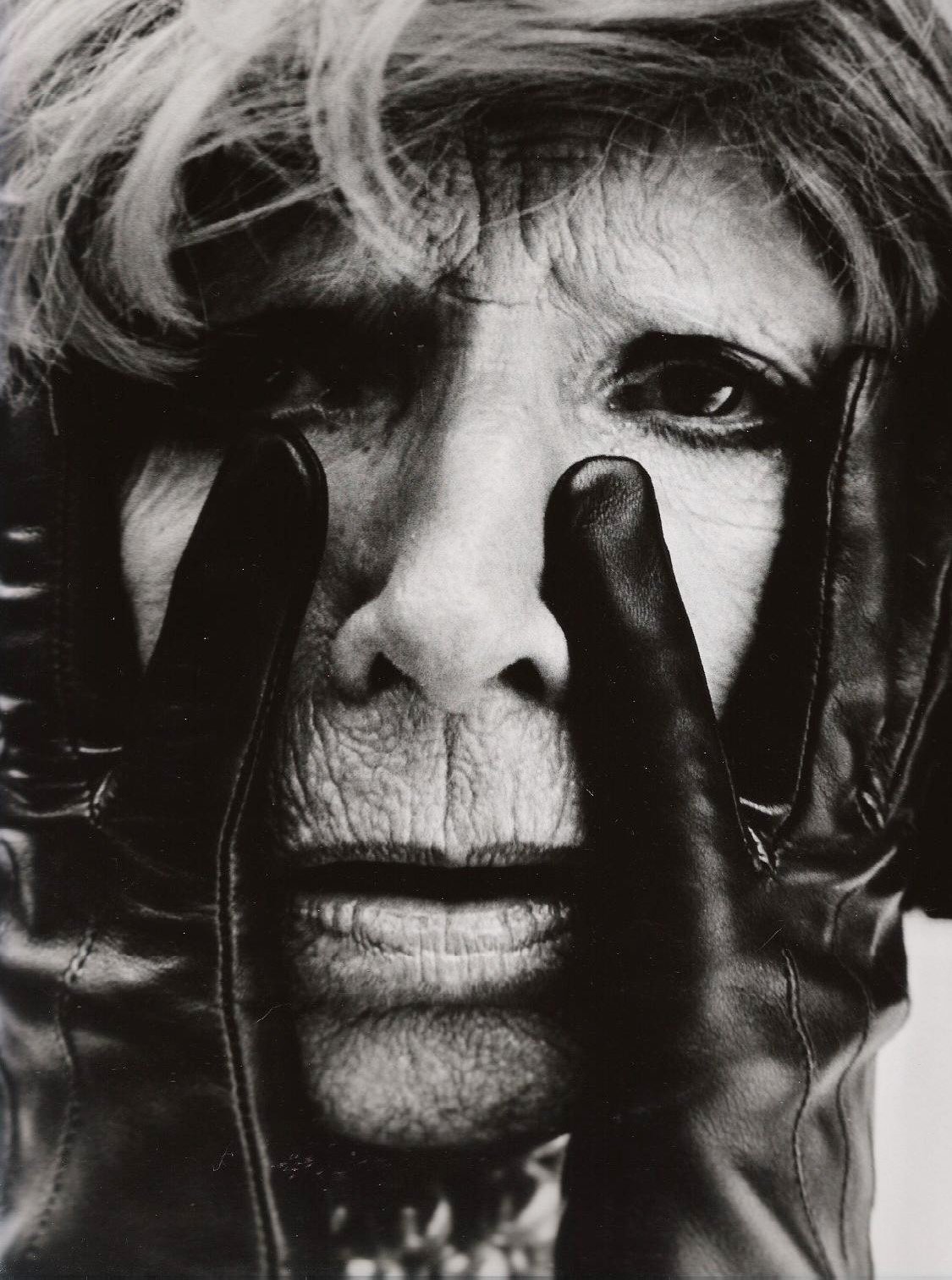 Close-up black-and-white photograph of an elderly woman with detailed facial wrinkles, wearing a leather jacket, with a gloved hand touching her face.