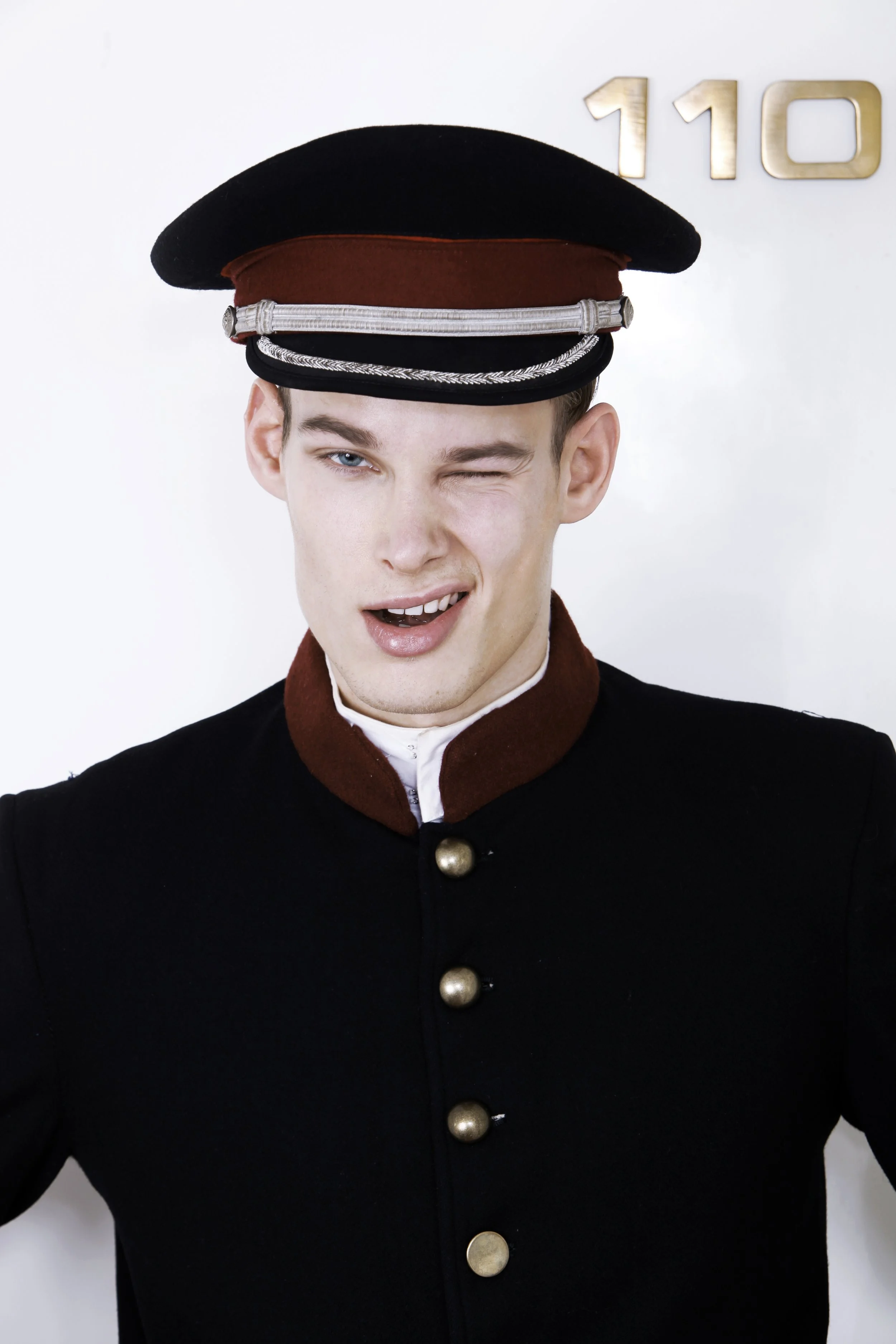 A person dressed in a vintage bellhop uniform, including a black hat with a red band and silver details, and a black jacket with large silver buttons, standing in front of a white wall with the number 110 in gold.