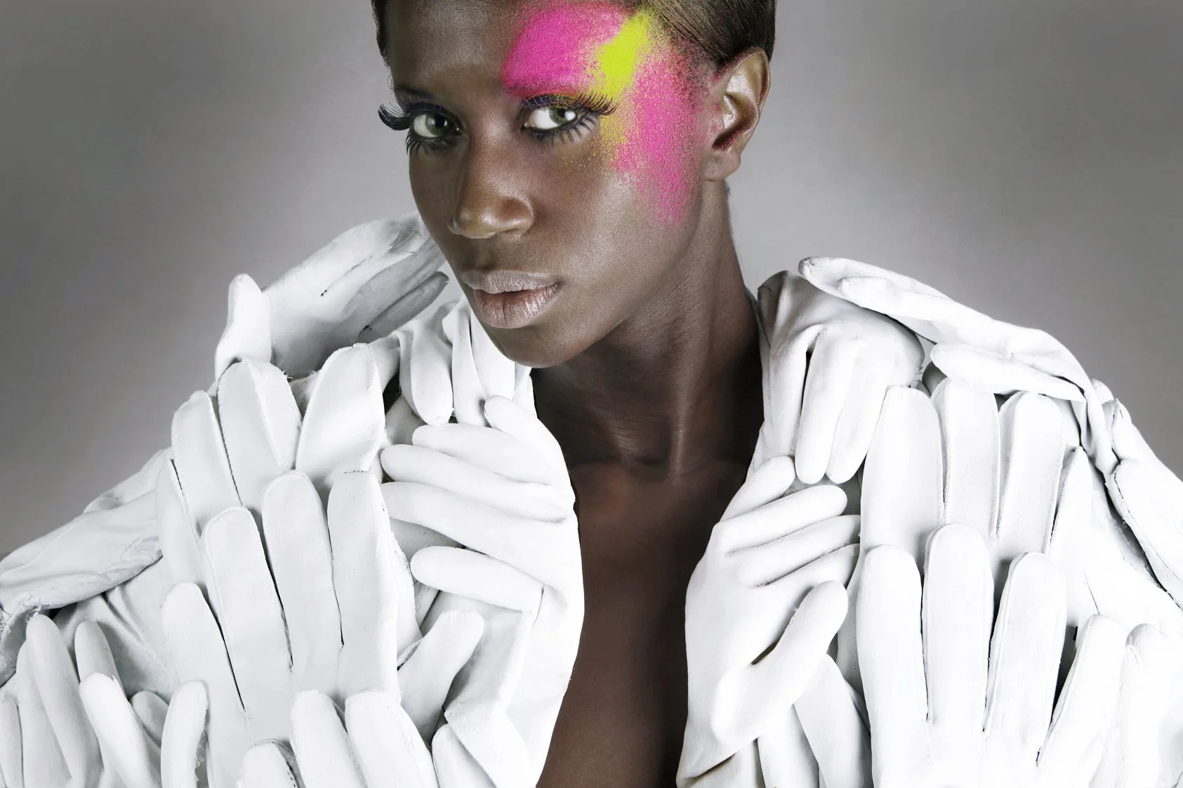 A woman with bright pink and yellow face paint, wearing white gloves and white clothing, looking at the camera.