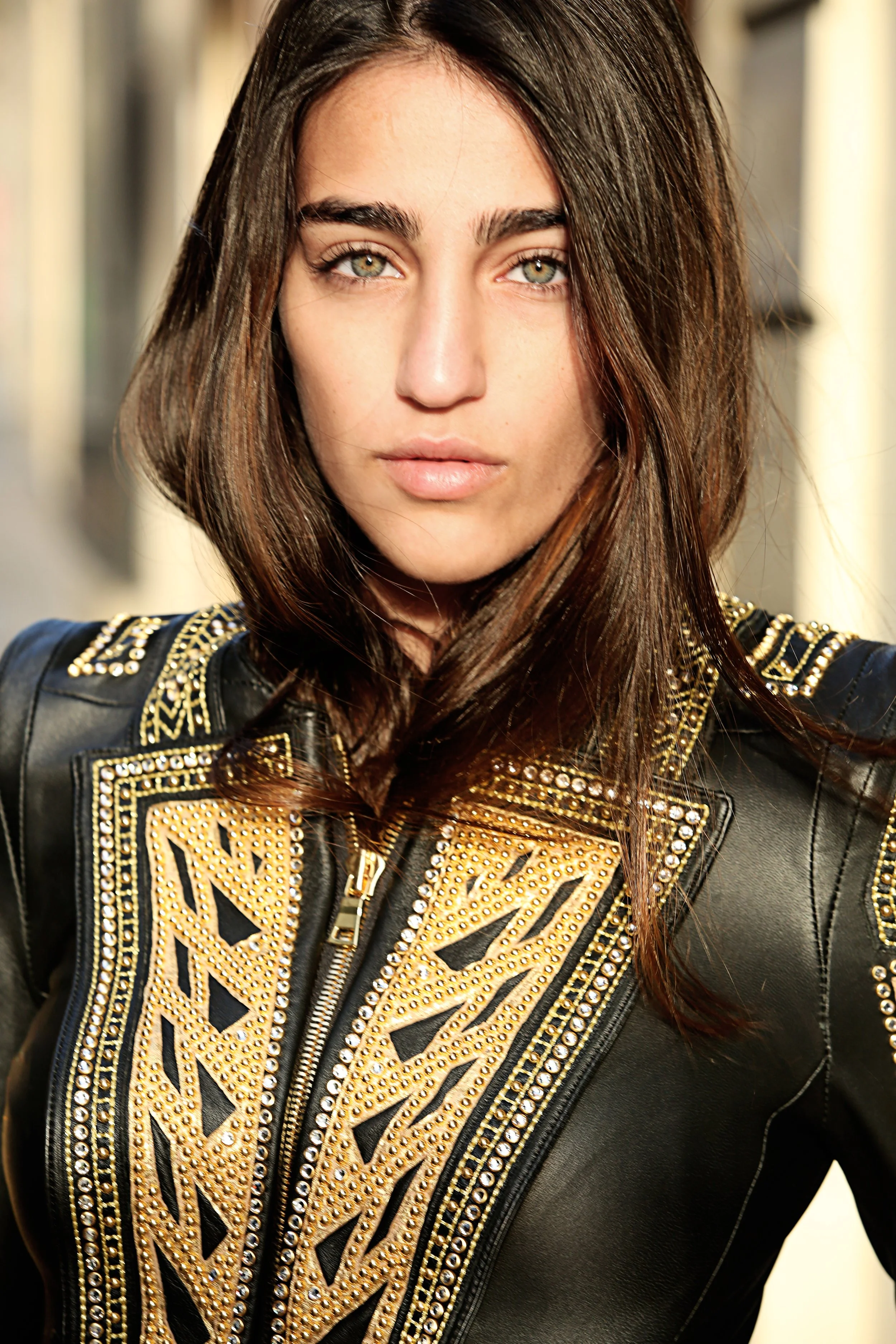 Close-up of a woman with long brown hair and hazel eyes, wearing a black leather jacket with gold embroidery and studs.