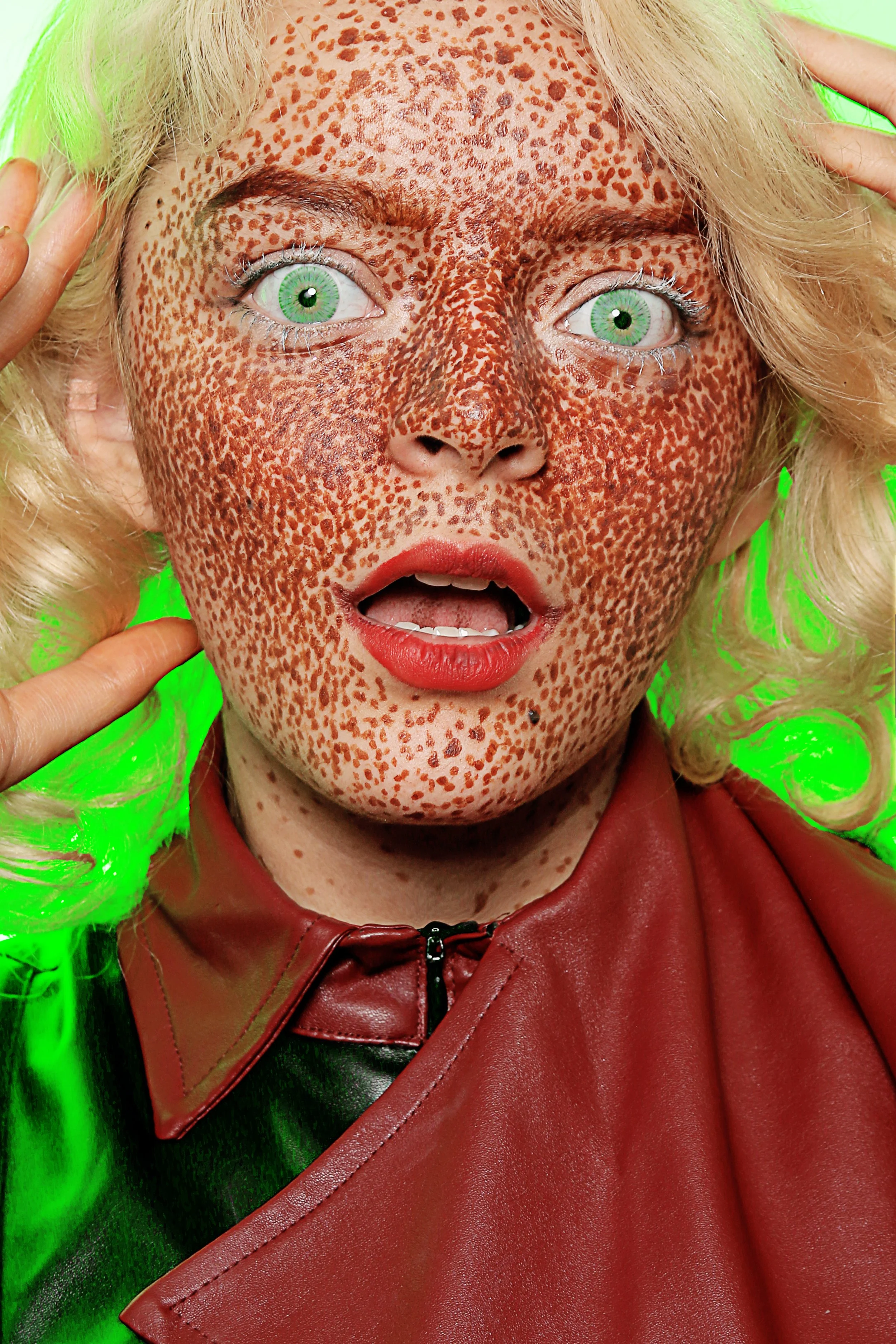 Close-up of a woman with green eyes, red lipstick, and a heavily freckled face. She has platinum blonde hair, wears a red leather jacket, and is holding her hair back with her hands against a bright green background.