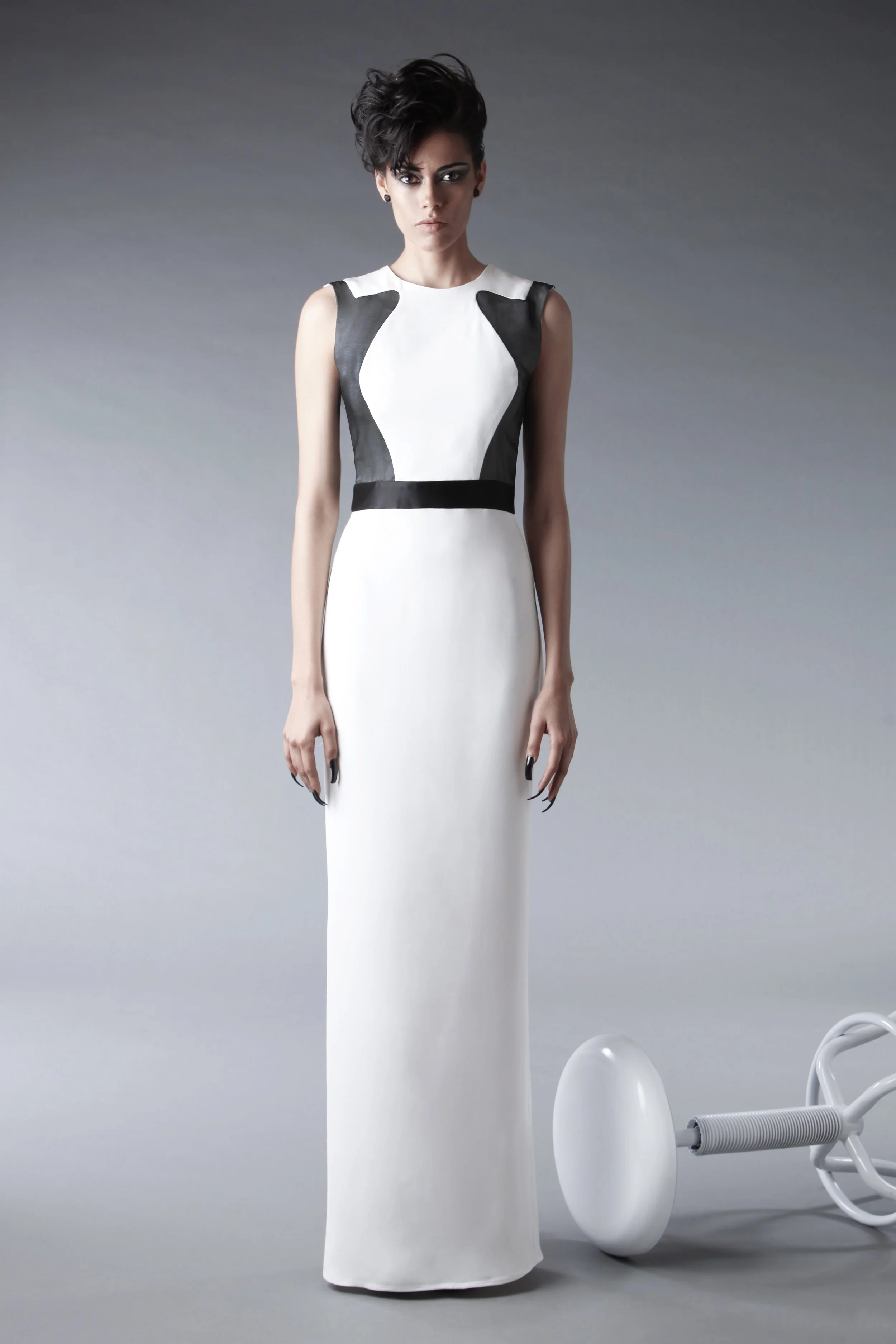 A woman in a sleeveless black and white dress stands in front of a gray background, with white modern chairs and objects to her right.