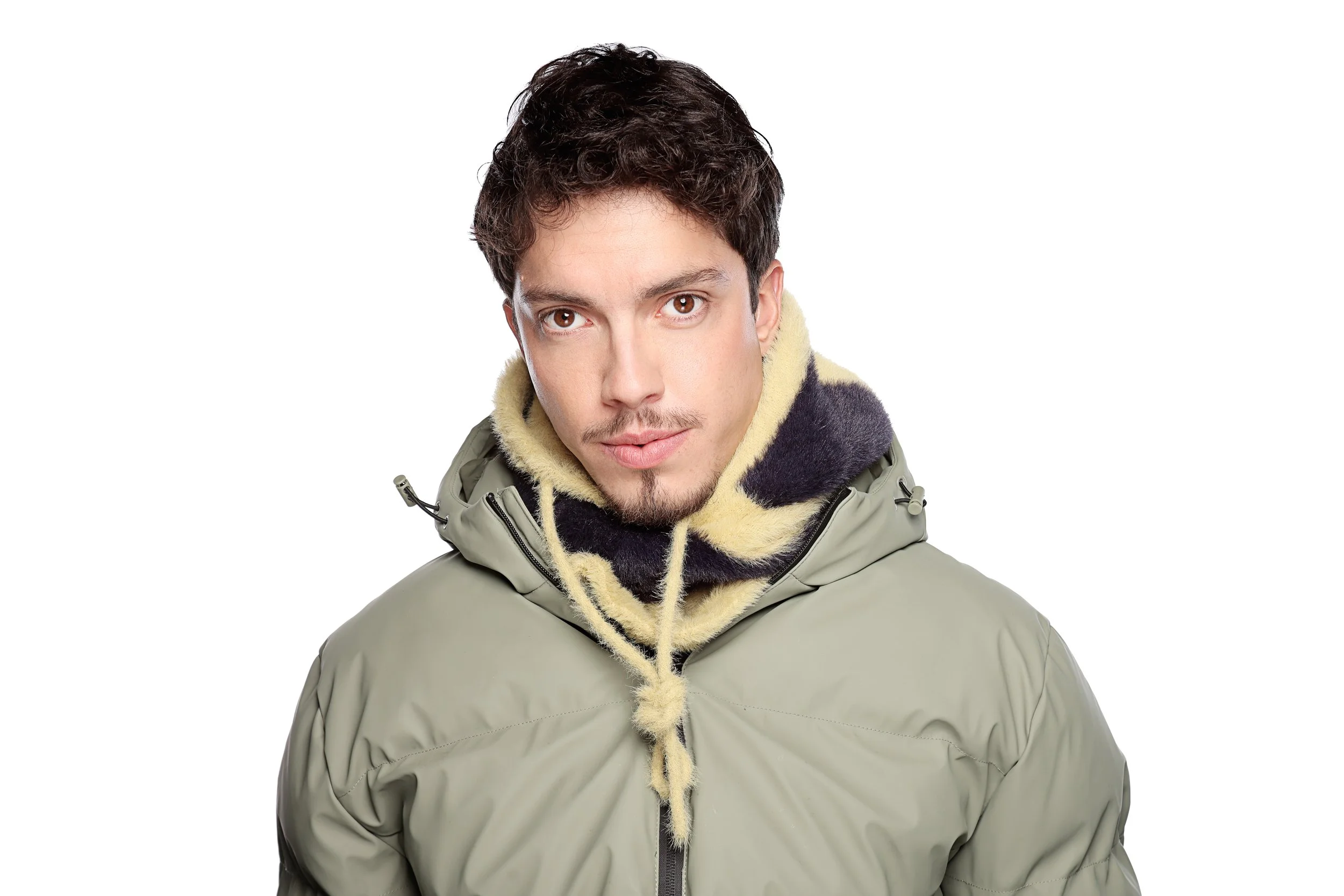 Young man with dark, curly hair and brown eyes wearing a light gray jacket with a hood lined with cream-colored faux fur, against a plain white background.