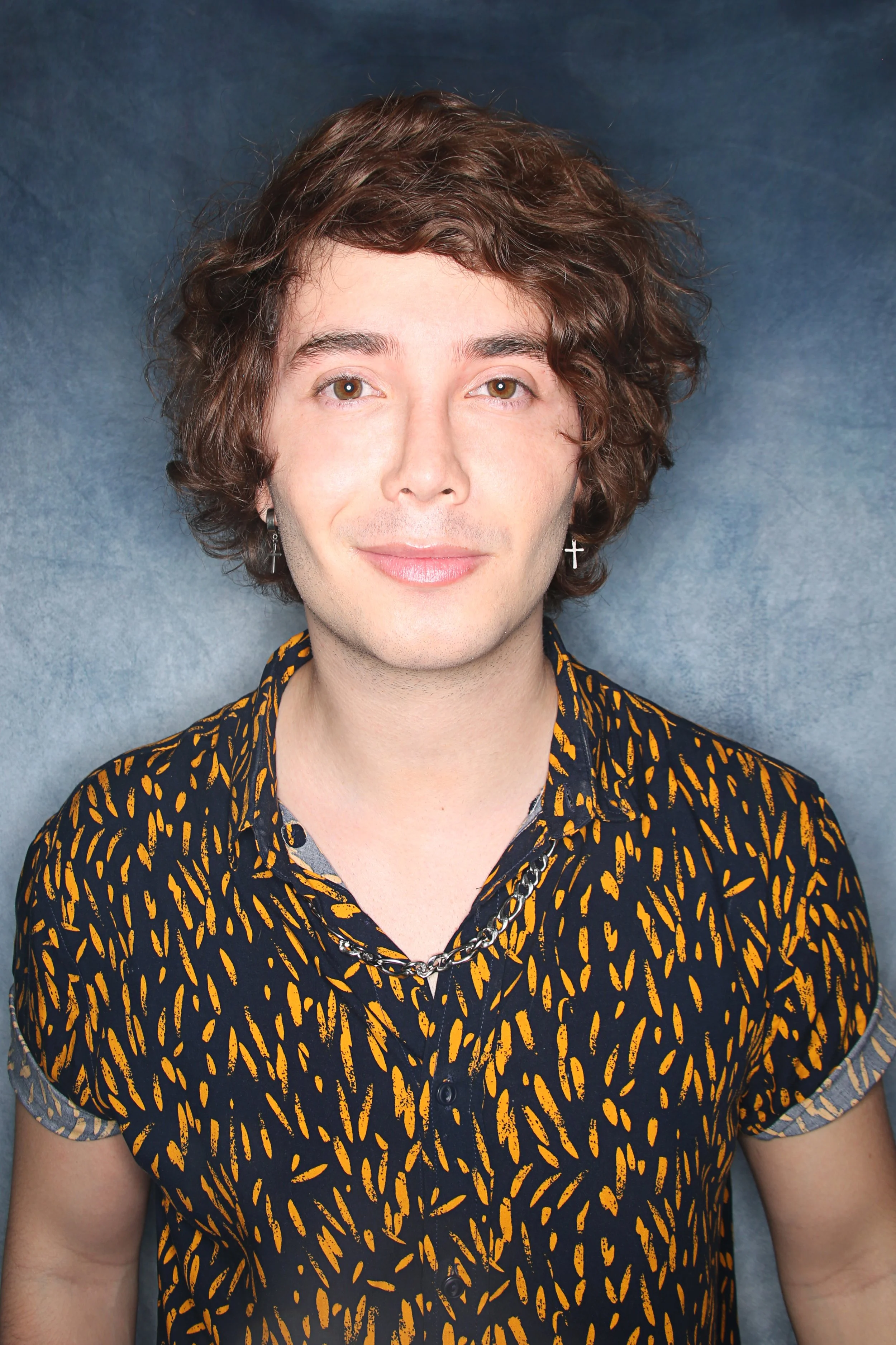 Portrait of a person with brown curly hair, wearing earrings, a patterned shirt with black and yellow designs, and a chain necklace, standing against a gray background.